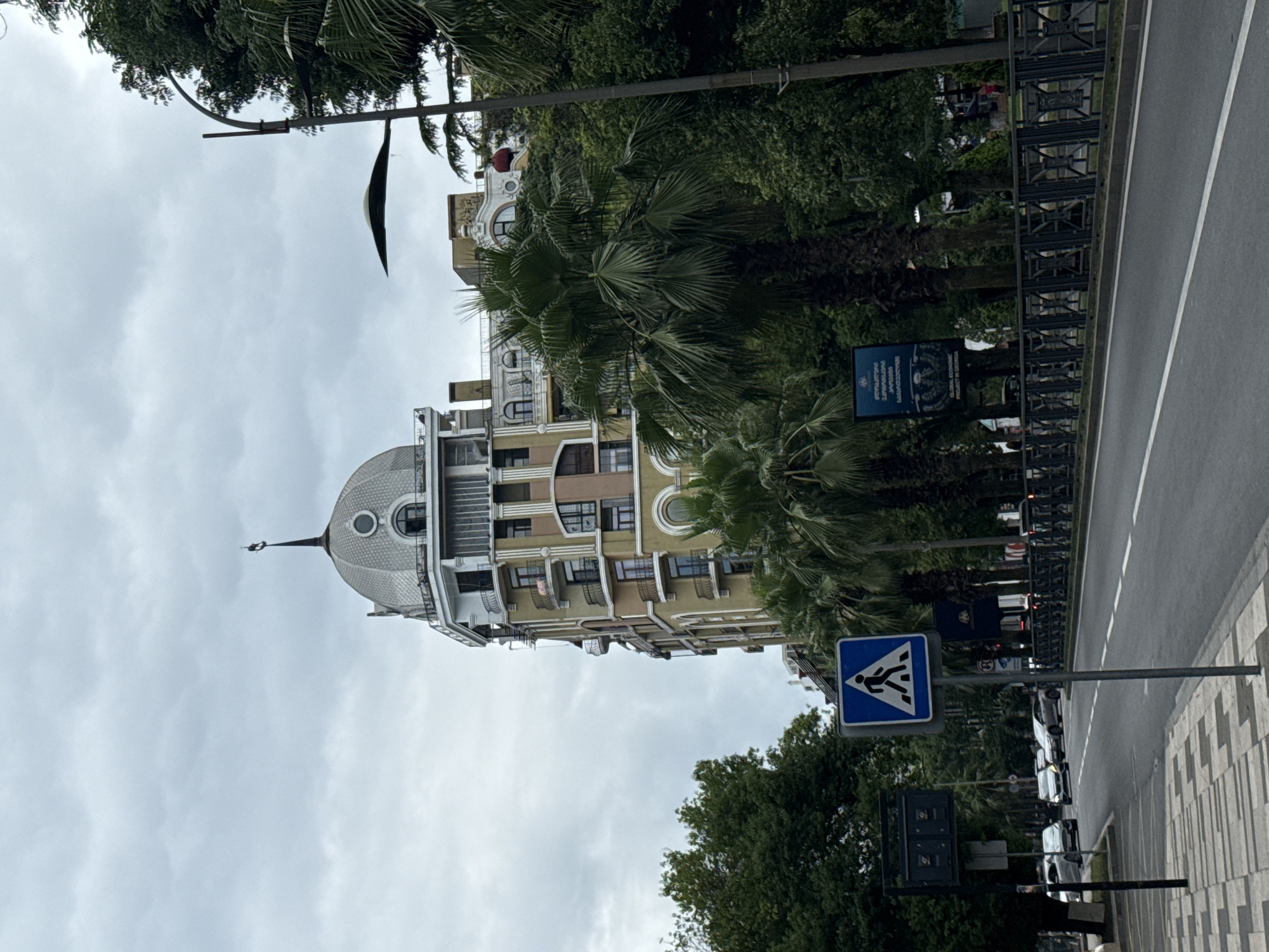 The image depicts a street scene in a city with a cloudy sky. A multi-story building with a dome-shaped roof and ornate balconies is visible on the right side of the street, surrounded by lush green trees, including palm trees. A pedestrian crossing sign is prominently displayed on the left side of the road. Cars are present on the asphalt road, which has white lane markings. The sidewalk is paved with a geometric pattern. A black metal fence runs along the side of the road, bordering a green space. There is a street lamp on the right. Two dark signs are mounted on poles on the left side of the road, one with what appears to be a digital display and another smaller sign below it. A large billboard with text and images is also visible behind the trees on the right. The overall impression is of an urban environment with a mix of modern architecture and natural elements.