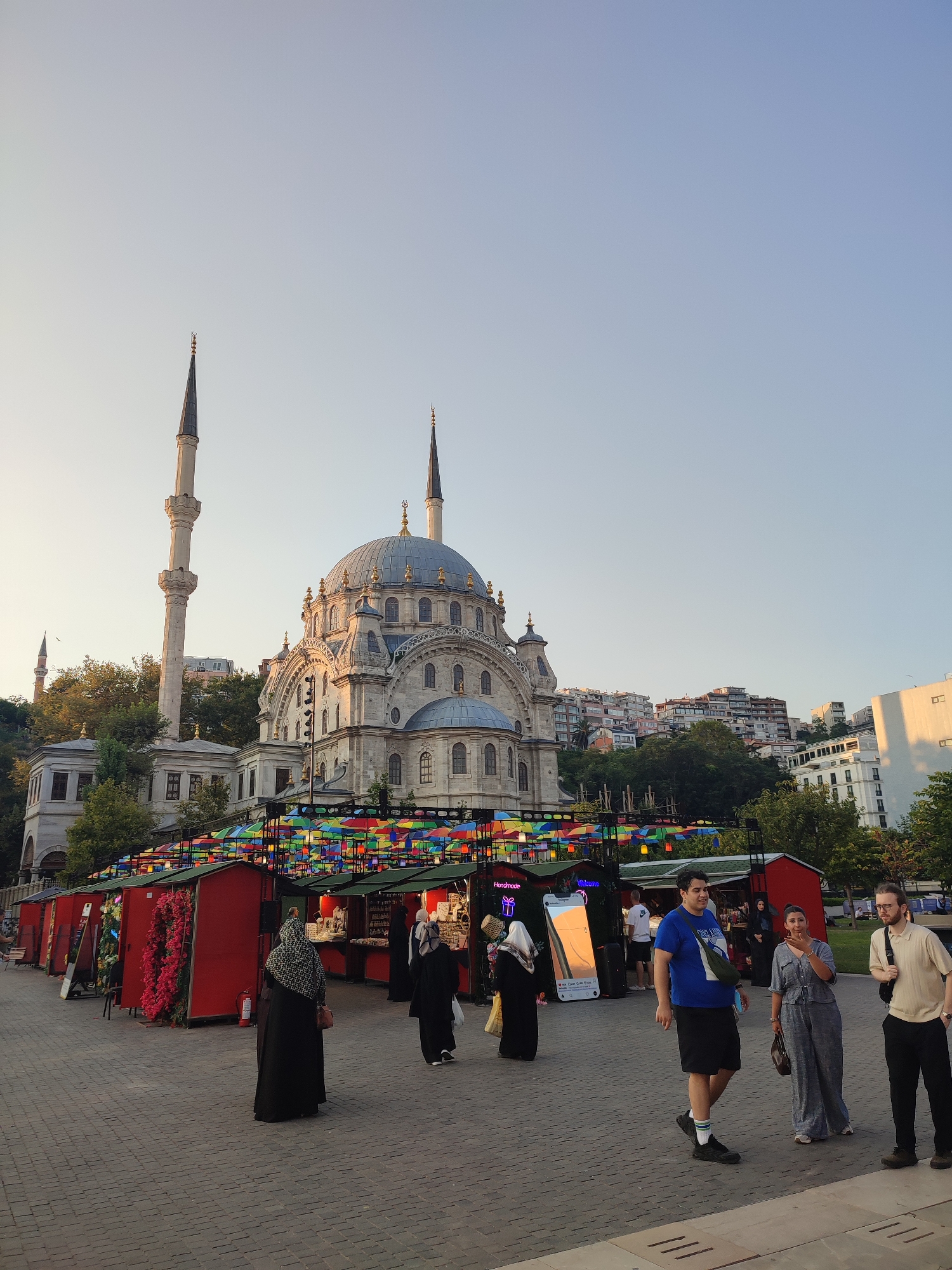 The image depicts a street scene in Beyoğlu, Turkey, featuring the exterior of a mosque with prominent minarets and a large dome. In front of the mosque, a row of red market stalls with colorful umbrella decorations are set up. Several people are visible in the foreground and mid-ground. Two women in dark Islamic attire are walking away from the camera. A man in a blue t-shirt and black shorts walks towards the camera, while a woman in a blue jumpsuit stands next to him. Another man in a light-colored shirt and black pants stands to the right, facing the camera. The ground is paved with cobblestones. The background shows a hillside with trees and residential buildings. The sky is clear and pale blue.