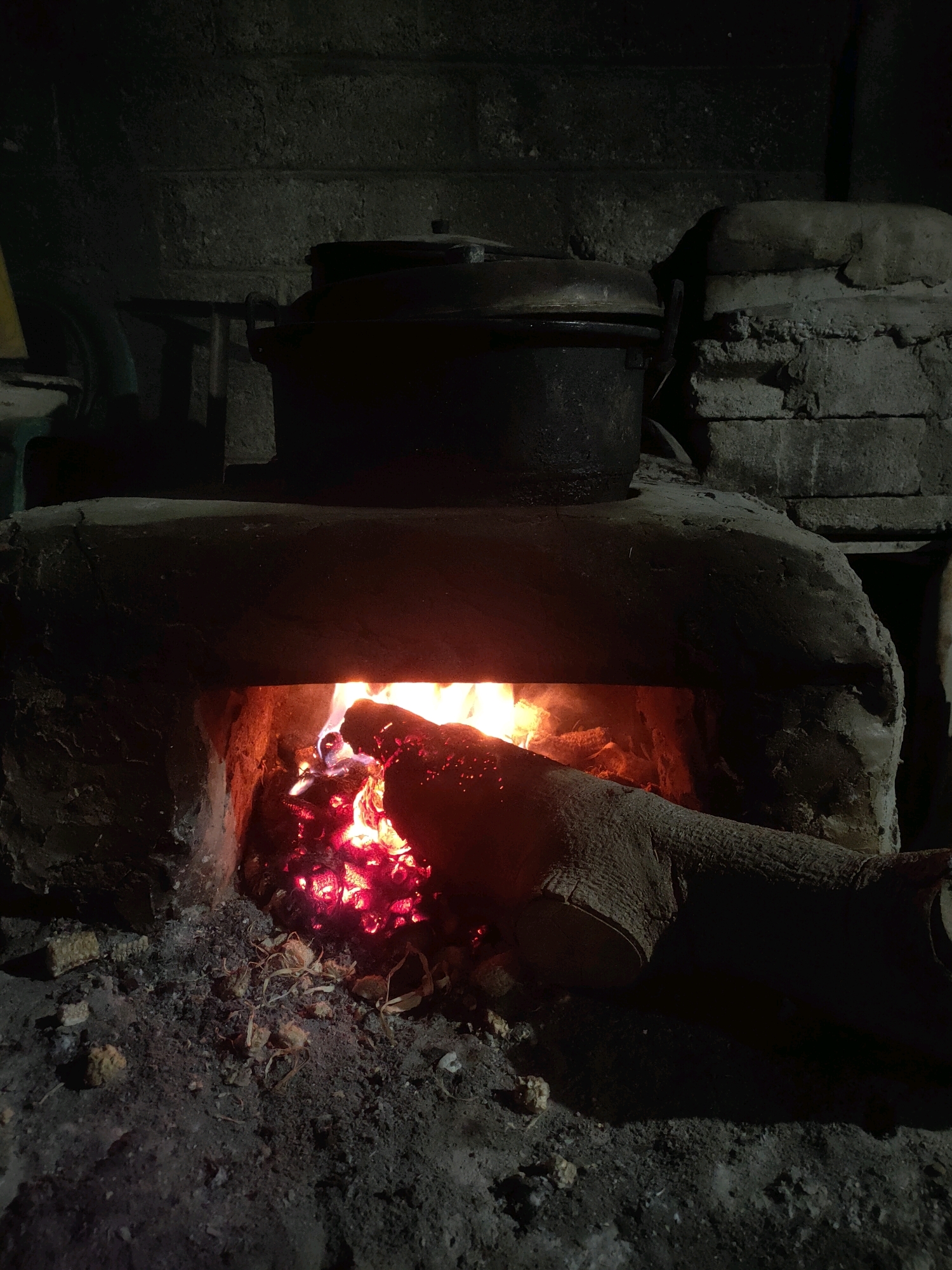An indoor shot of a rustic, brick oven with a fire burning inside is seen in the city of Plemahan, Indonesia. A large, dark pot sits on top of the oven. The oven's opening is illuminated by the glowing embers and flames from the fire, which are fueled by a large log positioned in the foreground. The surrounding area is dark and appears to be a workspace, with ash and debris scattered on the ground. The walls behind the oven are made of rough, dark brick.