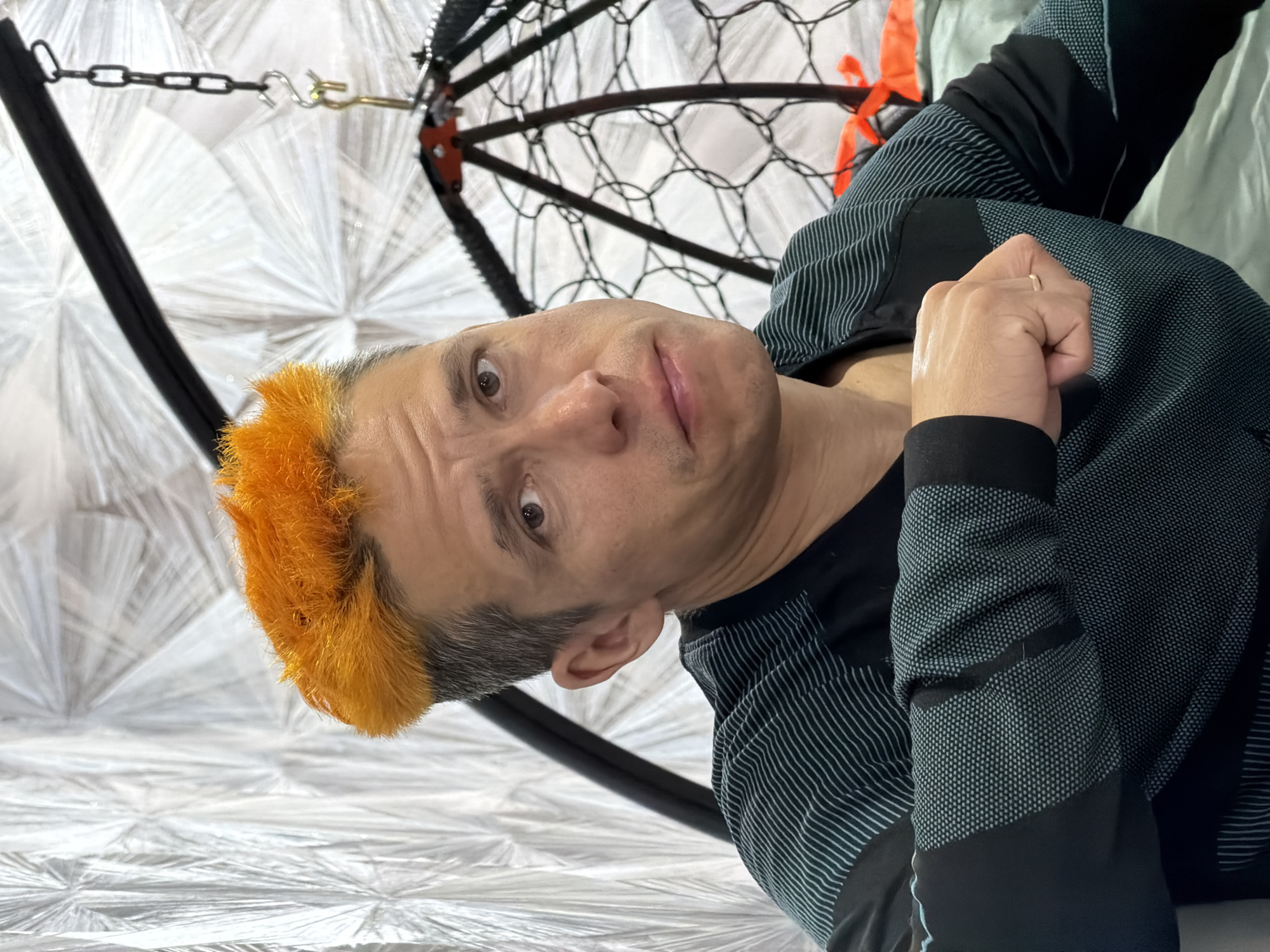 A medium-shot portrait depicts a person with short, spiky hair, vibrant orange at the front and transitioning to gray. The person looks directly at the camera with a neutral to slightly inquisitive expression. They are wearing a long-sleeved shirt that is black with patterns of thin teal/blue stripes on the sleeves and a dotted texture on the torso. The person's right hand, clenched into a fist and adorned with a thin gold band on the ring finger, rests against their body.
The background is dominated by a bright white, textured material, possibly fabric, featuring a distinctive radiating, folded pattern. To the right and above the person, parts of a dark, likely black, circular or semi-circular frame with a net-like interior are visible, suggesting it's a component of a hanging chair or swing. A metal chain with a hook and swivel connects to this overhead structure in the upper left of the frame. The overall setting appears to be indoors, potentially a recreational area or a uniquely decorated space. The lighting is diffused and even, offering no specific time-of-day cues. The image is taken at an angle, hinting at the person reclining. No visible text is present. The location context is Chaikovsky, Russia.