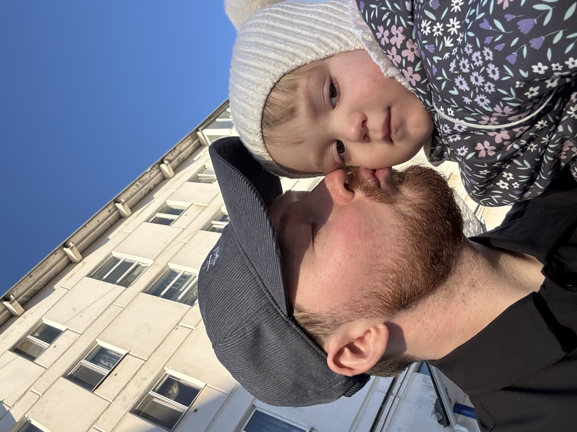 A close-up, tilted, low-angle shot shows a fair-skinned man with a reddish beard and mustache, wearing a gray corduroy baseball cap and a black shirt. He is nuzzling noses with a fair-skinned toddler wearing a white knit hat with a pom-pom and a floral-patterned garment. They are both looking at the camera. Behind them is a light-colored building with multiple windows and a blue sky. Based on visual cues of building architecture, the photo was likely taken in the city of Chaikovsky, Russia.