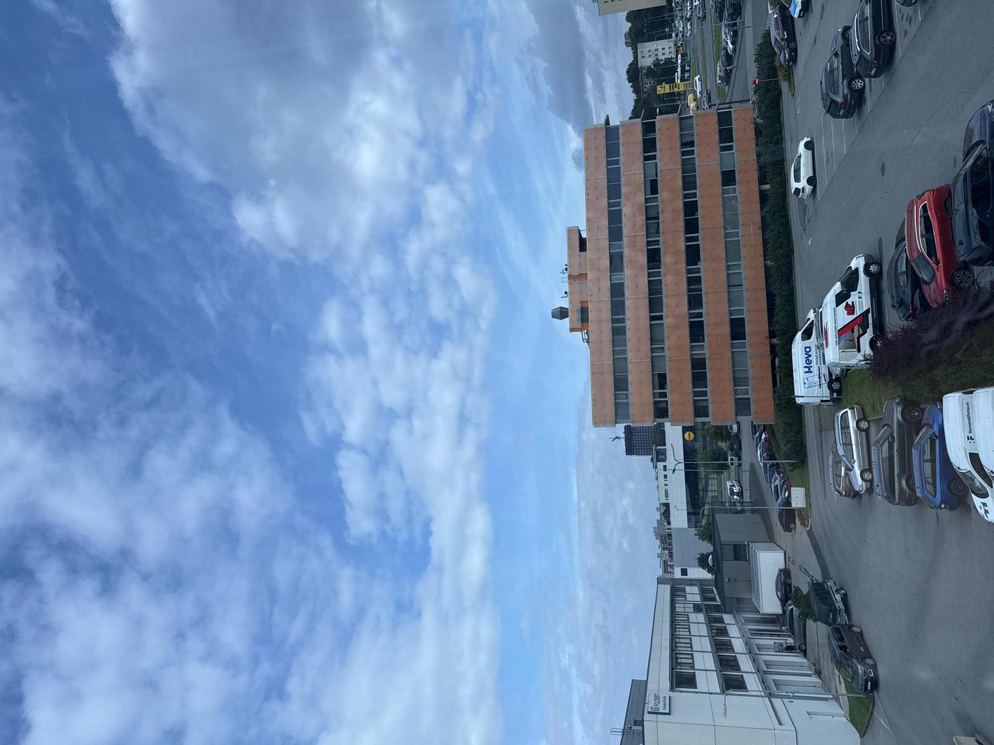 This is a high-angle, full shot of a parking lot and several buildings under a cloudy sky in Tallinn, Estonia. The buildings are modern, with a prominent orange-colored building in the mid-ground and a white building with a sign that reads "FOLDERIT" on the left. The parking lot is full of various cars, including sedans, SUVs, and vans. Some of the cars appear to be work vehicles, with branding such as "Heva" and "Remondifirma.ee" visible on them. The scene is daytime, with natural light illuminating the area. The sky is a mix of blue and white clouds, suggesting an overcast day. There are no people clearly visible in the image. The environment appears to be an industrial or business park.