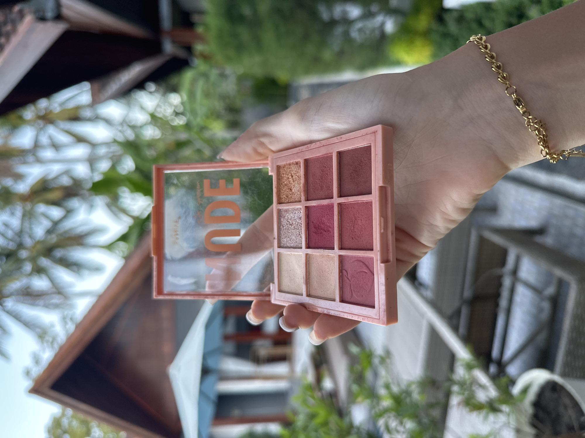 A hand wearing a gold heart-shaped bracelet holds an open, pink eyeshadow palette with nine different shades. The palette's cover is transparent with the word "NUDE" printed on it in orange. The background shows an outdoor setting with elements of a building's architecture, greenery, and hints of blue from an umbrella, suggesting a tropical or subtropical location. The architecture resembles Balinese-style structures. Considering the prompt includes "Nusa Penida, Indonesia," it is highly probable that this scene is set within that location.