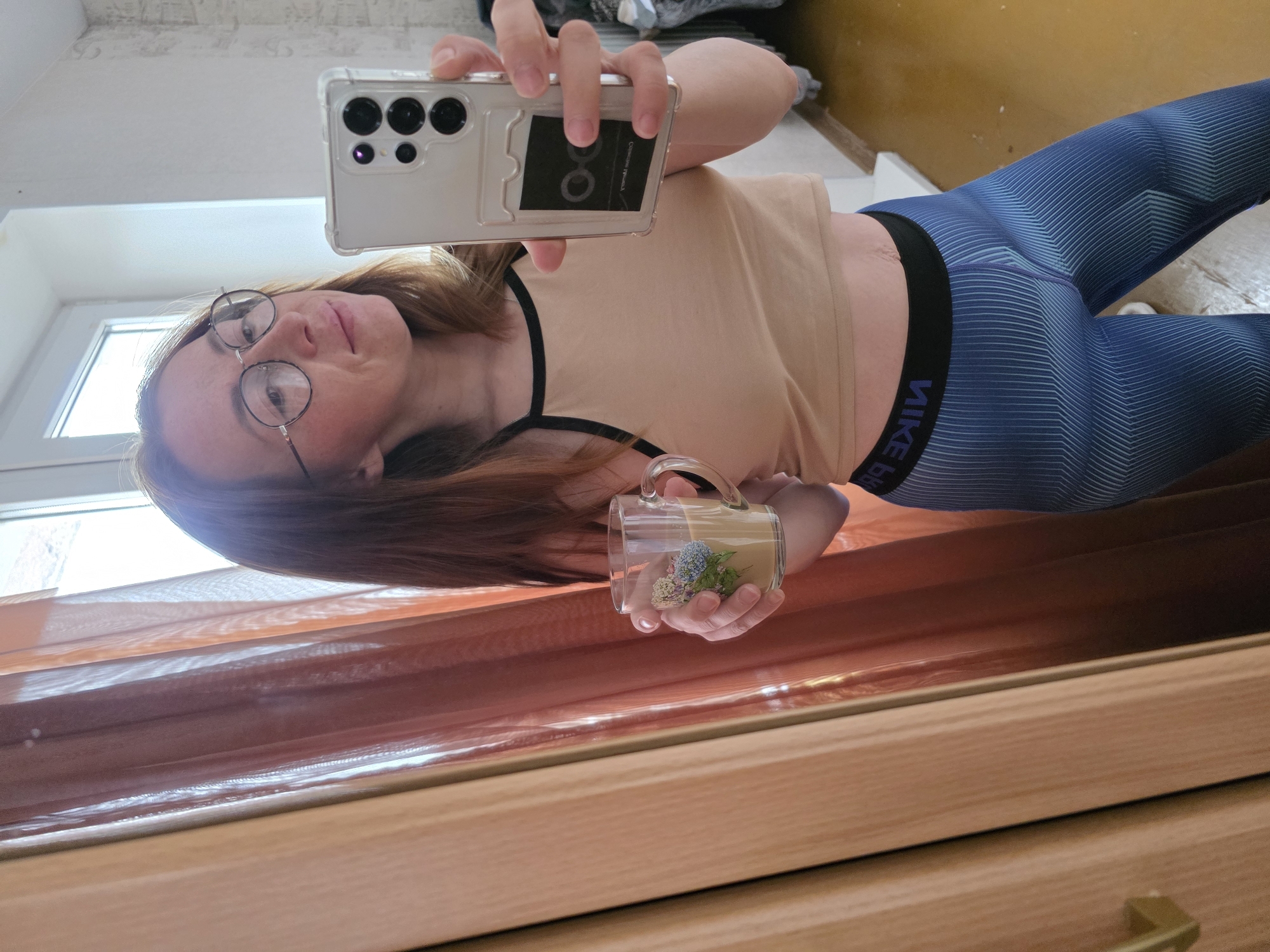 A woman is taking a selfie in front of a wooden dresser. She is wearing glasses, a tan crop top, and blue Nike leggings. She is holding a clear mug with a flower design in her left hand. In her right hand, she is holding a smartphone with a clear case. The background includes a window with natural light. The photo was taken indoors. While the location is mentioned to be in Chaikovsky, Russia, there is nothing in the image to independently verify this.