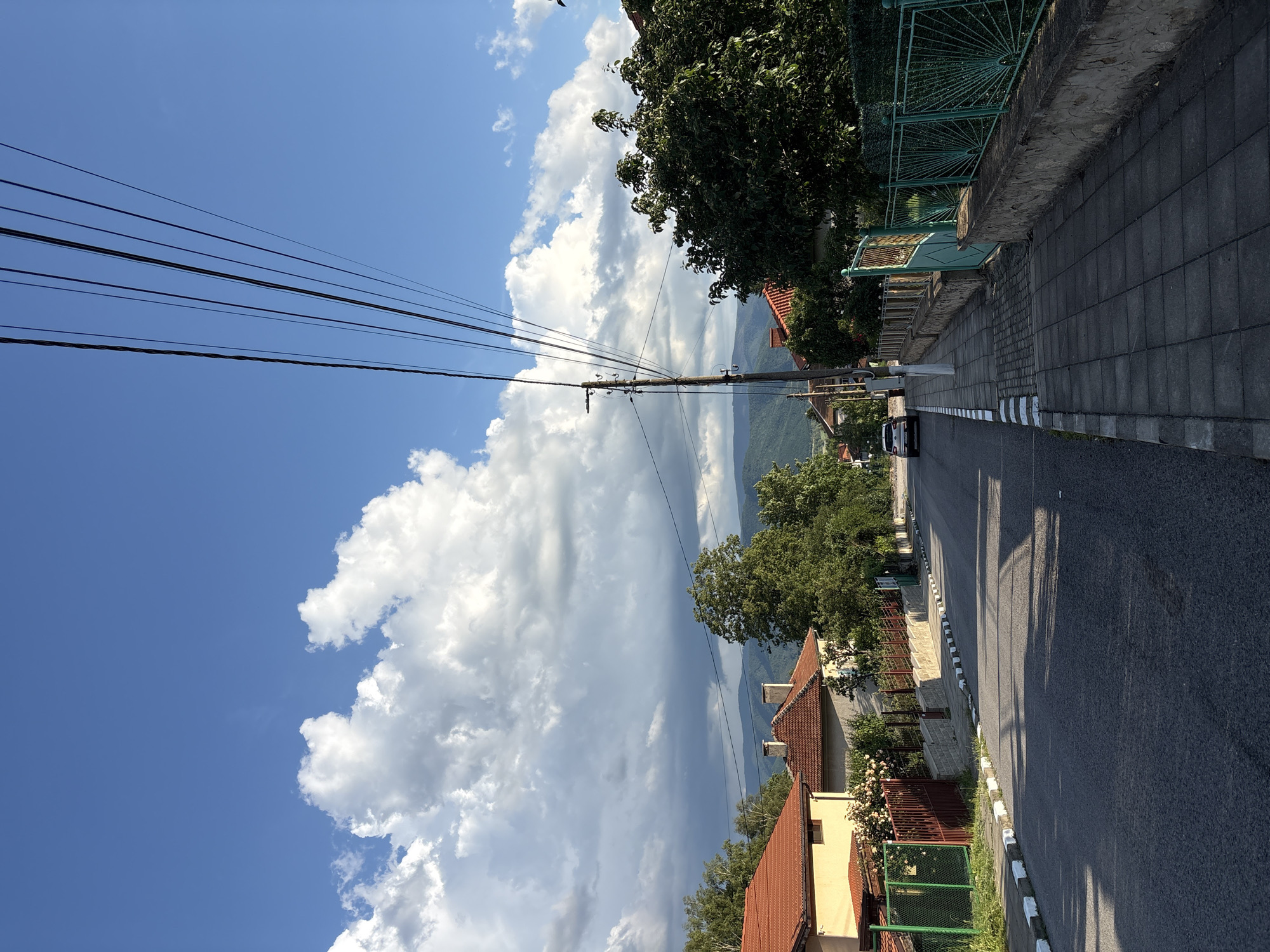This is a photograph of a street in the town of Anton, Bulgaria, under a partly cloudy blue sky. The road, paved with asphalt, runs diagonally from the lower left to the upper right of the frame, with a gray tiled sidewalk running parallel to it on the right. The sidewalk is bordered by a low stone wall topped with a green, ornate metal fence.
To the left of the road, houses with red-tiled roofs are visible, separated by green and brown fences. Trees and vegetation are present in front of and between the houses.
A wooden utility pole with numerous black wires stretches across the upper right portion of the image, its wires fanning out against the sky. In the distance, rolling hills covered in green foliage and trees form the backdrop. A white car is parked on the right side of the road, facing away from the camera. The scene is illuminated by natural daylight, casting shadows on the road and sidewalk.