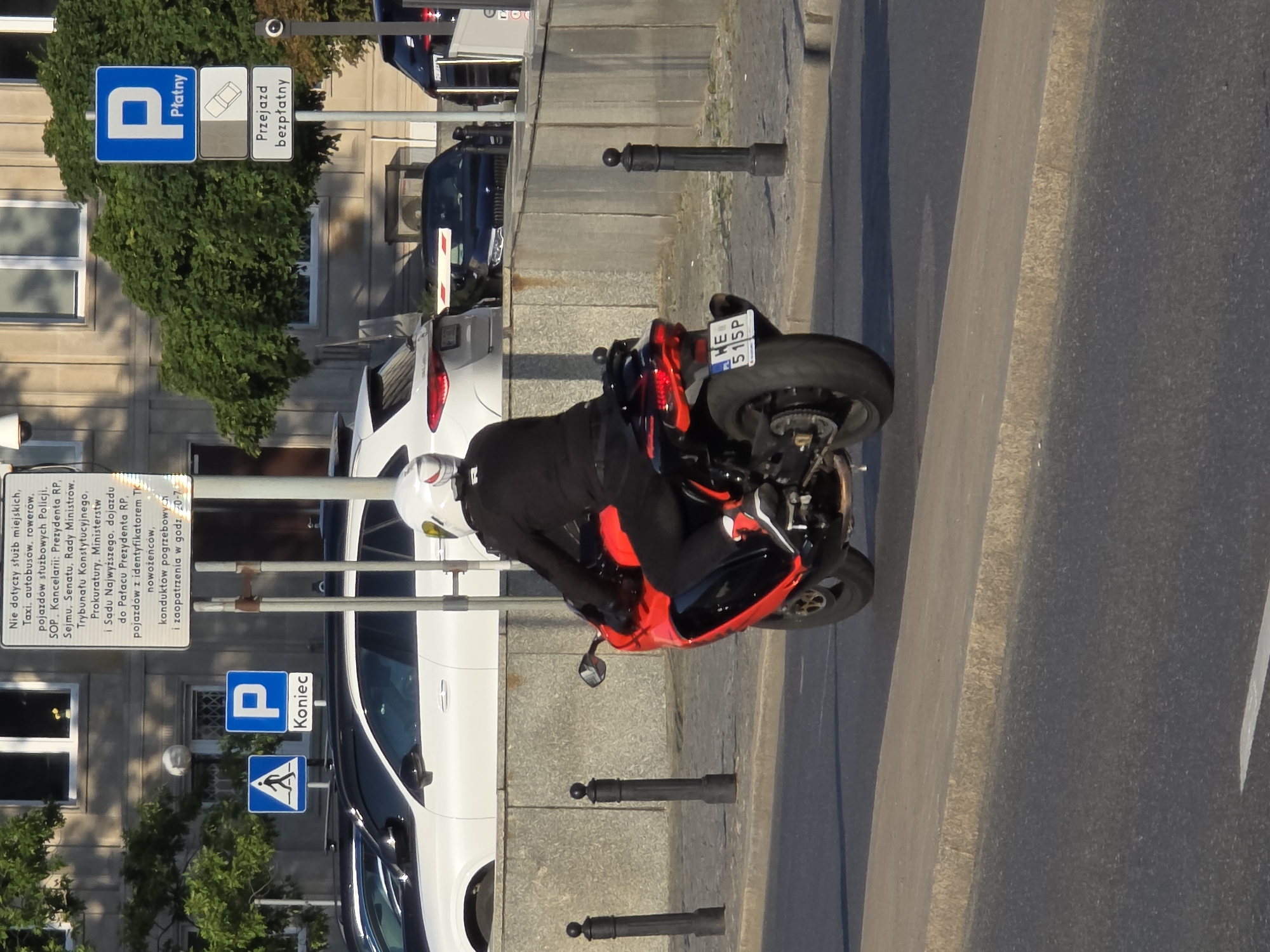 A motorcycle with a red and black body is being ridden by a person wearing a white helmet and a black jacket. The motorcycle has a license plate that reads "WE 515P". The motorcycle is on a paved road, next to a sidewalk and a stone wall. In the background, there are buildings and trees. There are also several traffic signs visible, including a "P" sign with "Koniec" underneath it, and another "P" sign with "Płatny" and a symbol of a car underneath it, followed by "Przejazd bezpłatny". These signs indicate a parking area where payment is required, but also free passage. The scene appears to be in an urban environment, possibly Warsaw, Poland, given the style of the license plate and the Polish text on the signs. The sun is shining, casting shadows.