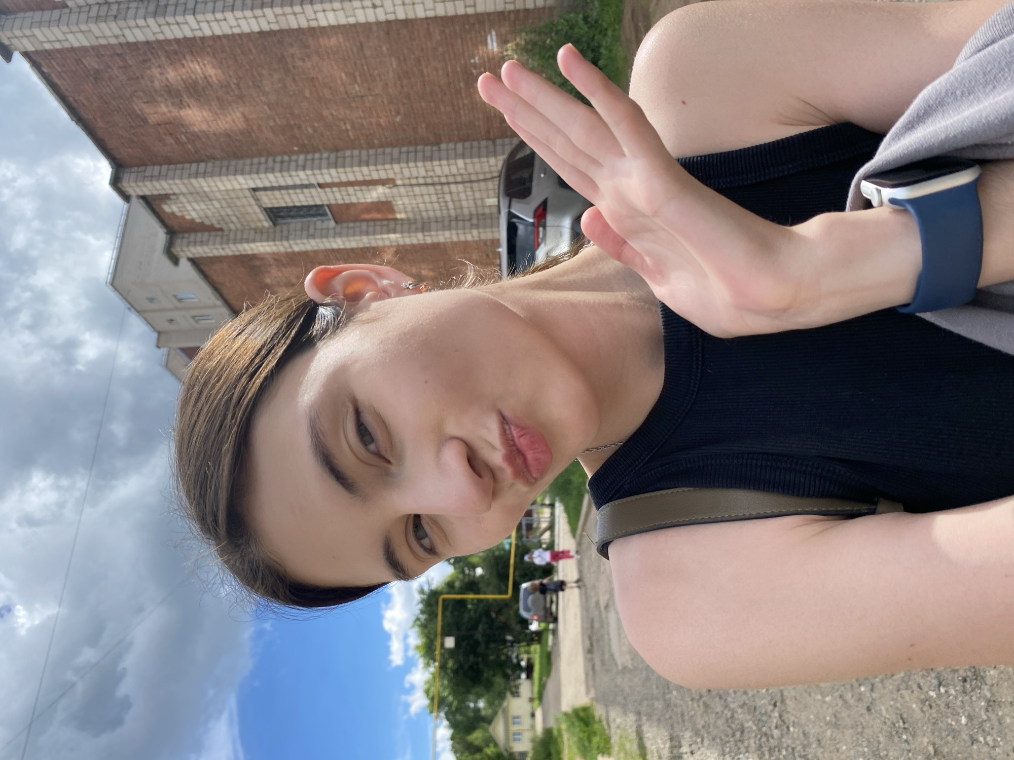 The media file is a portrait-oriented selfie taken outdoors. The main subject is a young person with brown hair, wearing a black top and a grey garment draped over their shoulder. They are making a "kiss face" with their lips pursed. Their right hand is held up, palm facing the camera, in what could be interpreted as a "stop" gesture. A blue-strapped smartwatch is visible on their left wrist.
The background suggests an urban or suburban setting in Votkinsk, Russia. A brick building with white trim is prominent on the left, with a cloudy sky above. To the right, a street or pathway is visible, with a parked car, some trees, and residential buildings in the distance. The lighting indicates it is daytime, likely sunny with some clouds. The overall mood appears casual and lighthearted. There is no visible text in the media file.