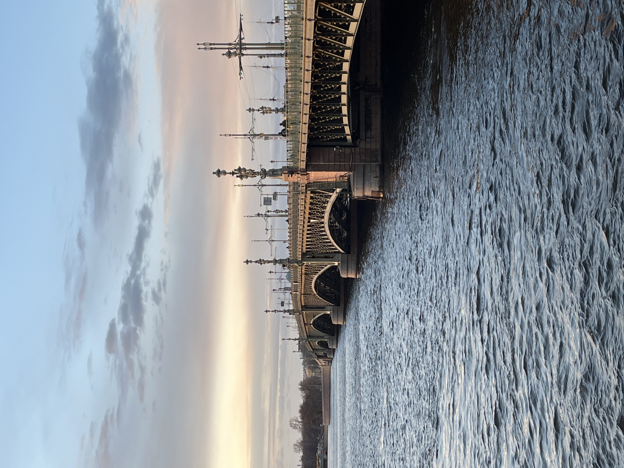 The image captures the Troitsky Bridge in Saint Petersburg, Russia, seen from a low-angle perspective. The bridge spans a body of water, presumably the Neva River, which occupies the right half of the frame and exhibits a rippled surface. The bridge's structure, including its arches and ornate lampposts, is visible along the left side of the image. The sky, which occupies the upper portion of the frame, is filled with clouds and varying shades of blue and gray. The image is rotated 90 degrees counter-clockwise.