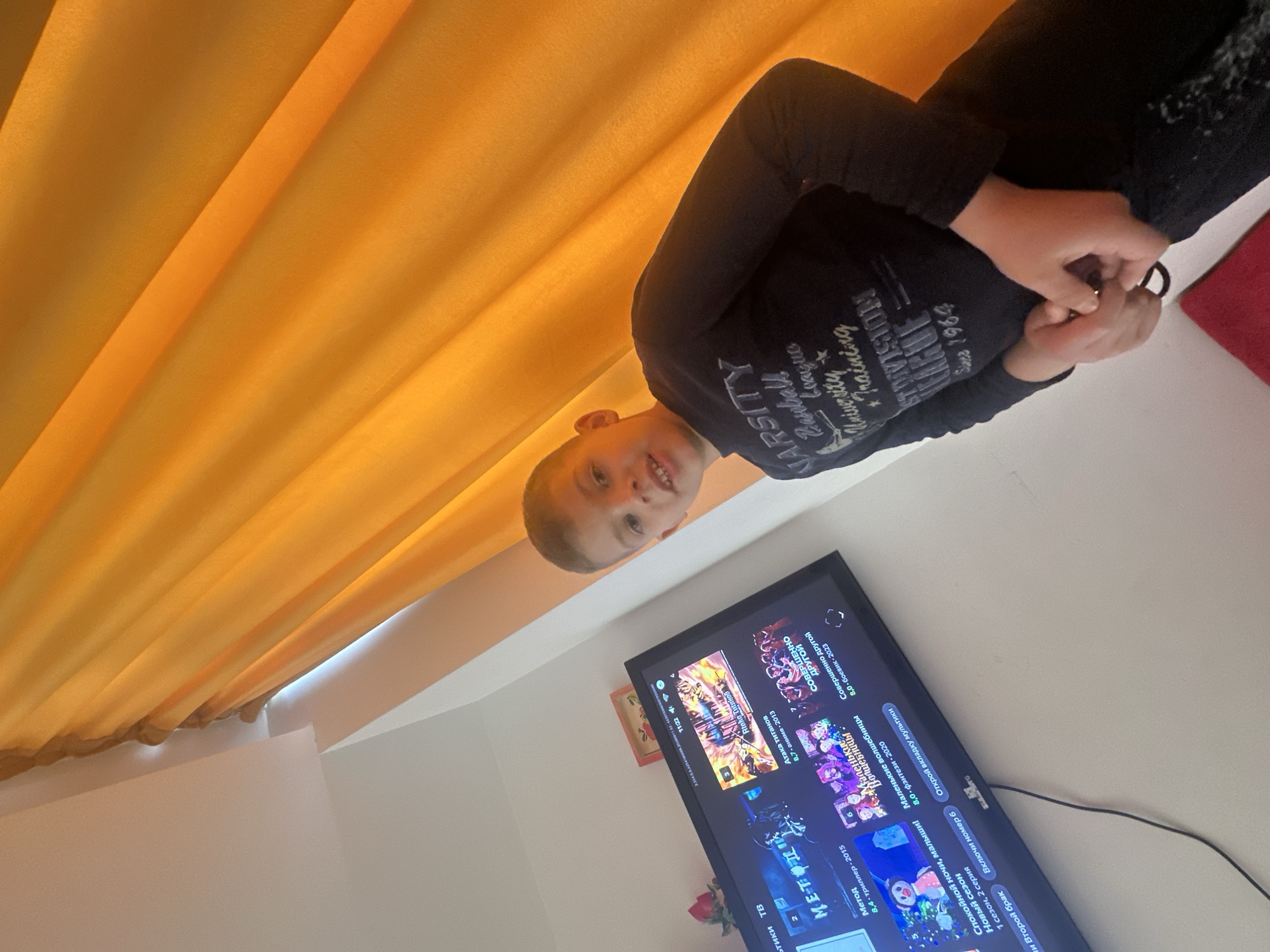 This is a vertical, candid shot of a young boy sitting in front of a television. The boy is wearing a dark long-sleeved shirt with white lettering that reads "VARSITY BASEBALL LEAGUE UNIVERSITY TRAINING ST DIVISION SINCE 1964". He is looking towards the camera with a smile. The television screen is displaying a menu with various show titles and ratings in Russian. The background consists of bright orange curtains and a white wall. The lighting suggests it is daytime. The boy appears relaxed and happy, engaged with the content on the screen.