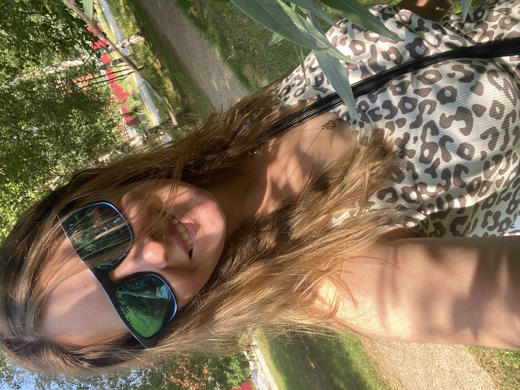 A woman with long, light brown hair and wearing sunglasses smiles at the camera. She is outdoors, possibly in a park or a wooded area, with trees and a path visible in the background. She is wearing a leopard-print top and a gold necklace. The sun is casting shadows on her face and arm.