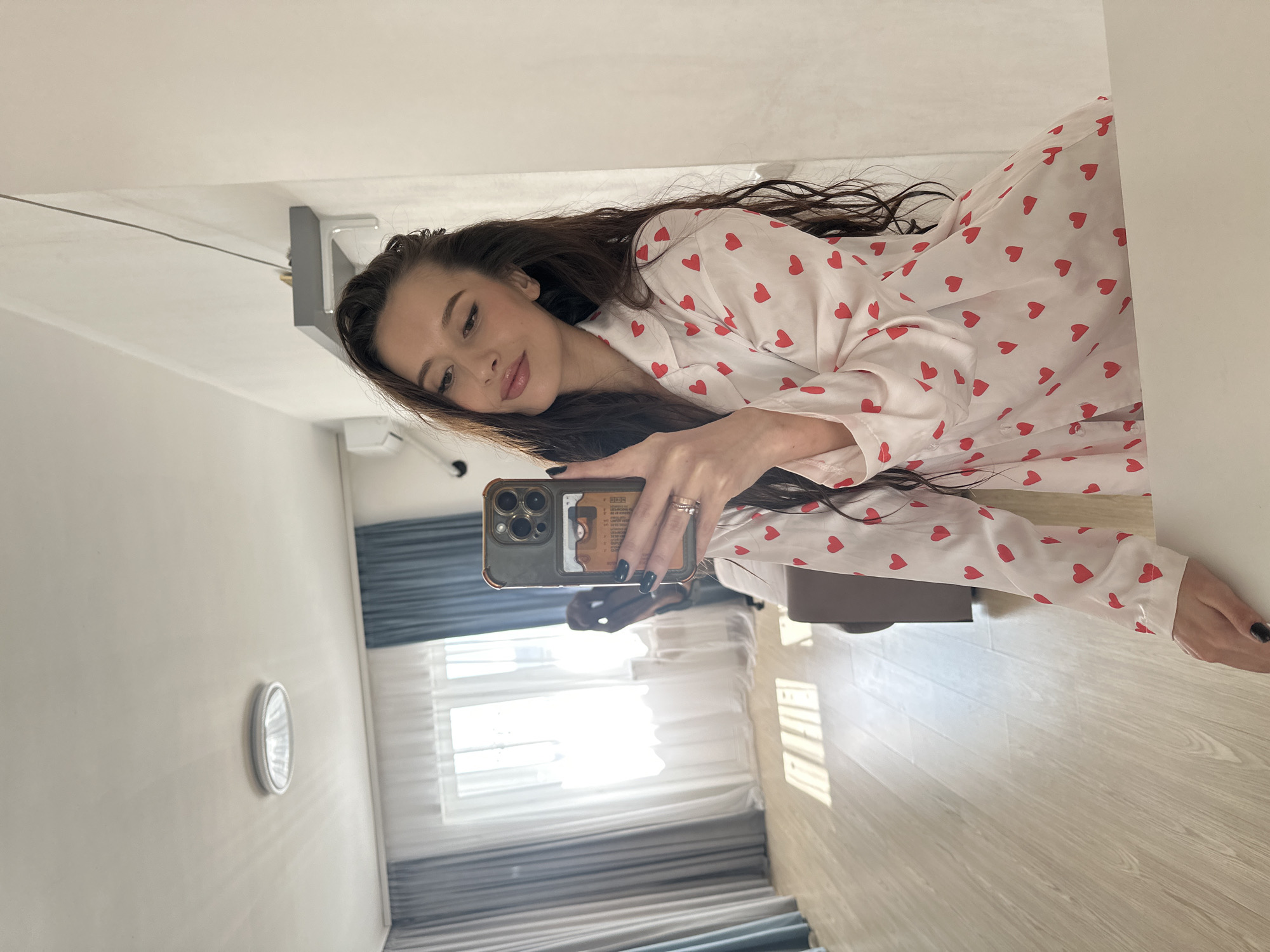 A woman is taking a selfie in what appears to be an indoor setting. She is wearing a long-sleeved pajama top adorned with red hearts. She is holding a smartphone with a distinctive case, which is casting a reflection of a window with curtains behind her. The room is illuminated by natural light streaming through the window. There is a wall-mounted light fixture visible on the left. The flooring consists of light-colored wooden planks. Although it's not possible to pinpoint the exact location within Yekaterinburg, Russia, based solely on the image, the interior setting suggests a residential or hotel environment.