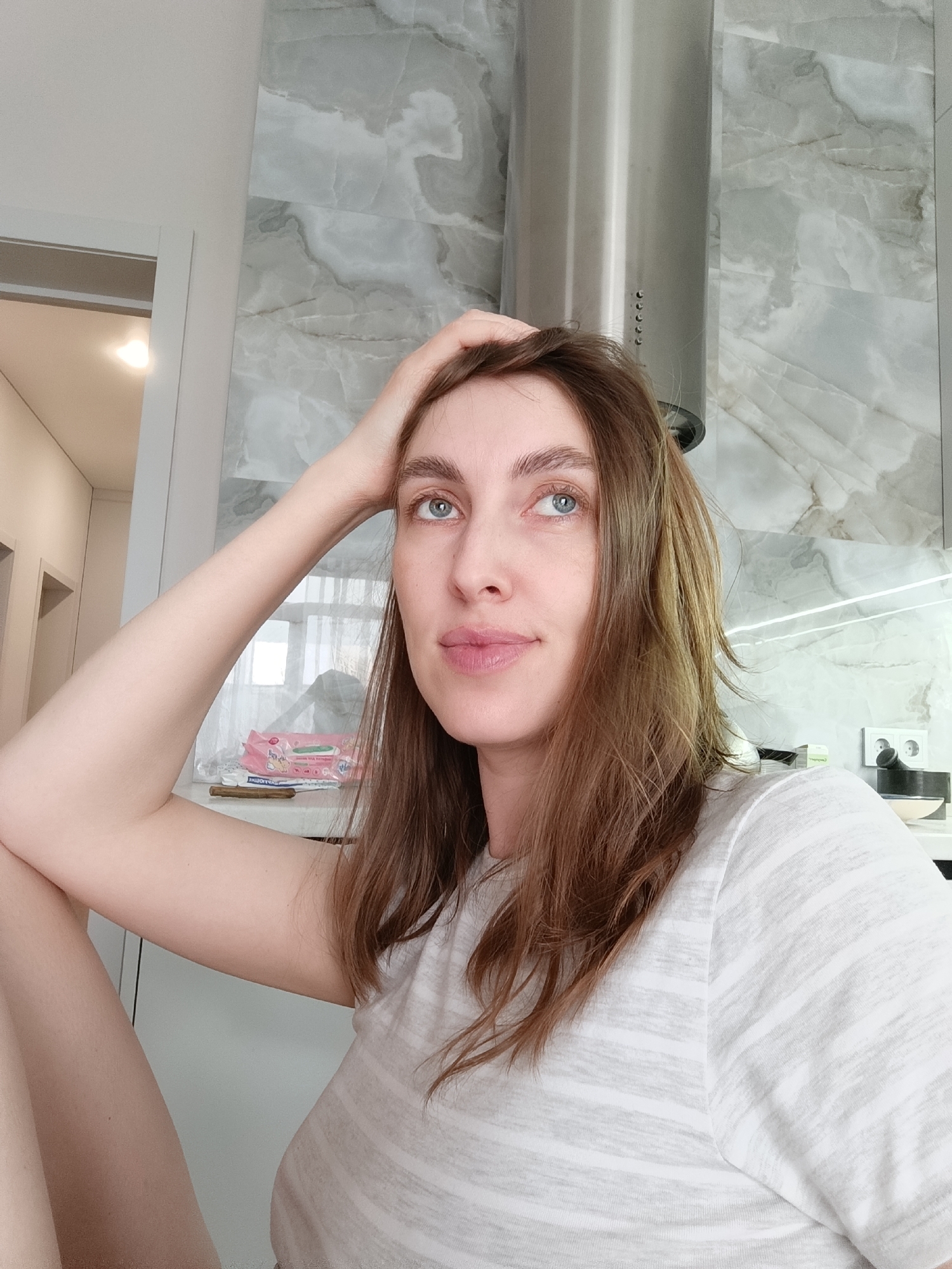 A young woman with long, medium-brown hair and striking blue eyes is depicted in a thoughtful, upward gaze, her right hand gently resting on her head. Her lips are slightly parted, conveying a pensive expression. She is wearing a light-colored t-shirt with subtle horizontal stripes.
The setting appears to be a modern kitchen or an open-plan living space in a residence in Chaikovsky, Russia. The background features a wall adorned with large, light grey and white marble-patterned tiles. A sleek stainless steel range hood is visible in the upper right. Below it, a white countertop is illuminated by a linear LED light strip.
On the countertop, to the left of the woman's arm, rests a pink packet of wet wipes. Partially visible Cyrillic text on the packaging includes "УЛЫ" (likely part of "Улыбка," meaning "Smile") and "БЕЗ СПИРТА" ("alcohol-free"). Beside the wipes, a small, dark, cylindrical wooden object is present. Further right on the counter, a white electrical outlet with two sockets is mounted on the tiled wall, next to a dark, small mortar and pestle. To the far left, a doorway leads to another room, where an interior light is on. The scene is bright and suggests daytime indoors, capturing a quiet, reflective moment.