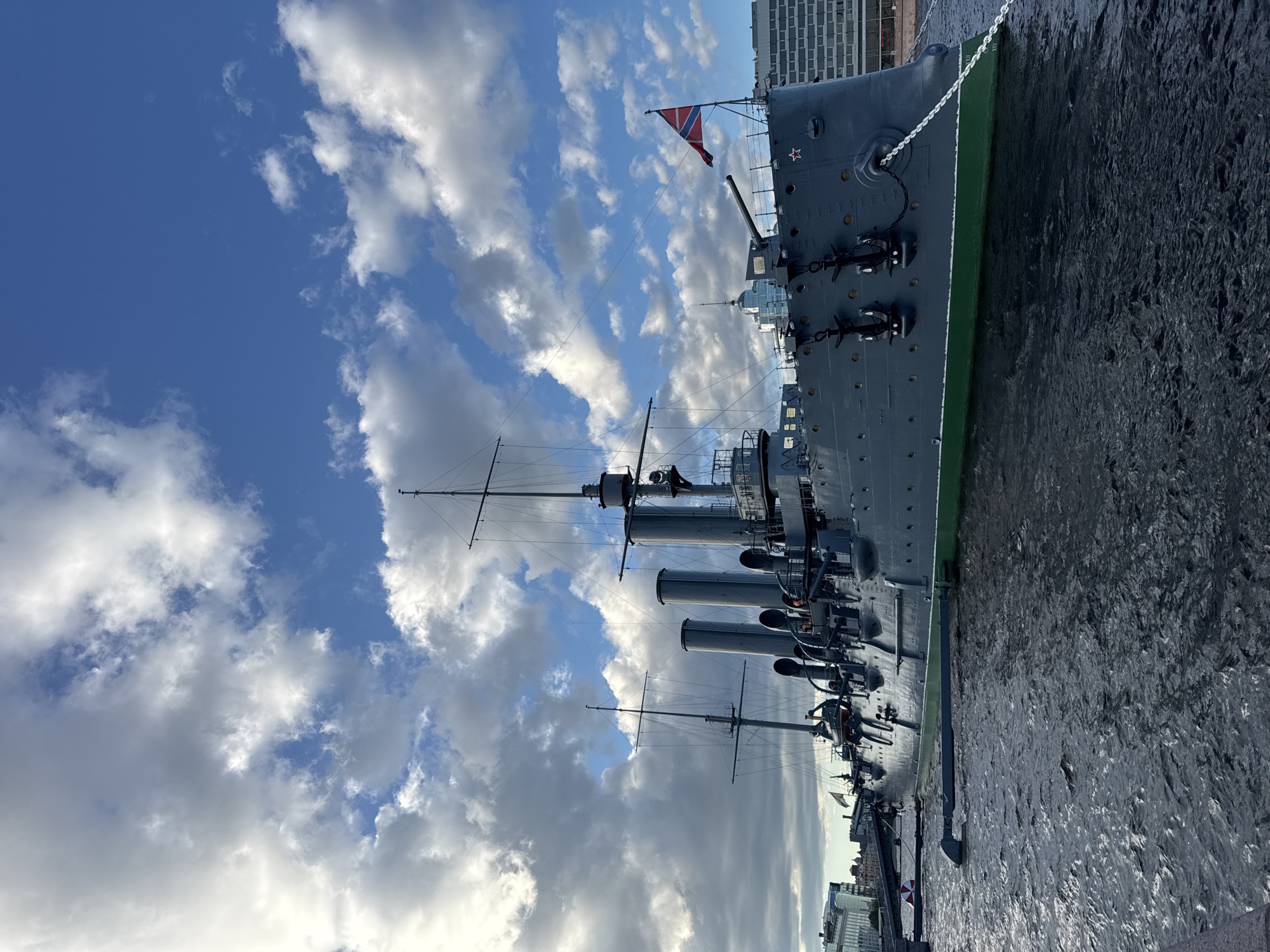 The image captures a wide-angle view of the cruiser Aurora, a historic ship docked on the Neva River in Saint Petersburg, Russia. The ship, painted in dark gray with a green waterline, is the central subject. It features multiple smokestacks, masts, and visible cannons. A Russian flag flies from the stern. The ship is moored in the water, with dark, choppy waves lapping against its hull. In the background, modern buildings and the cloudy sky are visible. The overall impression is that of a sunny day with dramatic clouds.