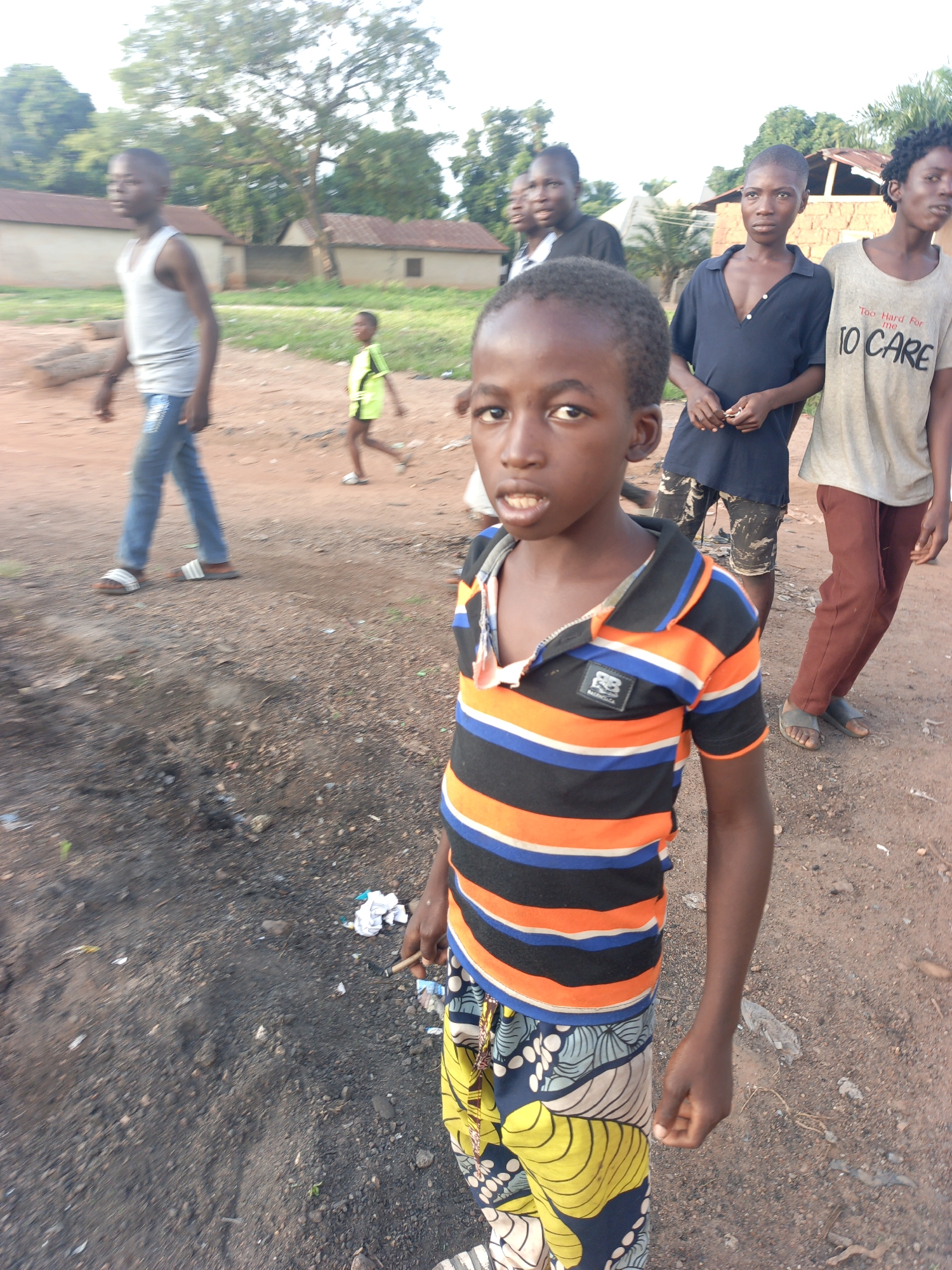 In Akwanga, Nigeria, a group of boys are gathered on a dirt road in front of low-rise buildings. The central figure, a young boy with a shaved head, wears a colorful striped shirt and patterned pants. To his right, another boy in a dark blue shirt and camouflage shorts stands with his hands clasped. Further back, a boy in a grey t-shirt with text and a girl in a green shirt are also present. The ground is covered in dirt and small stones, with some scattered debris. The environment appears to be a residential area with modest structures and vegetation in the background.
