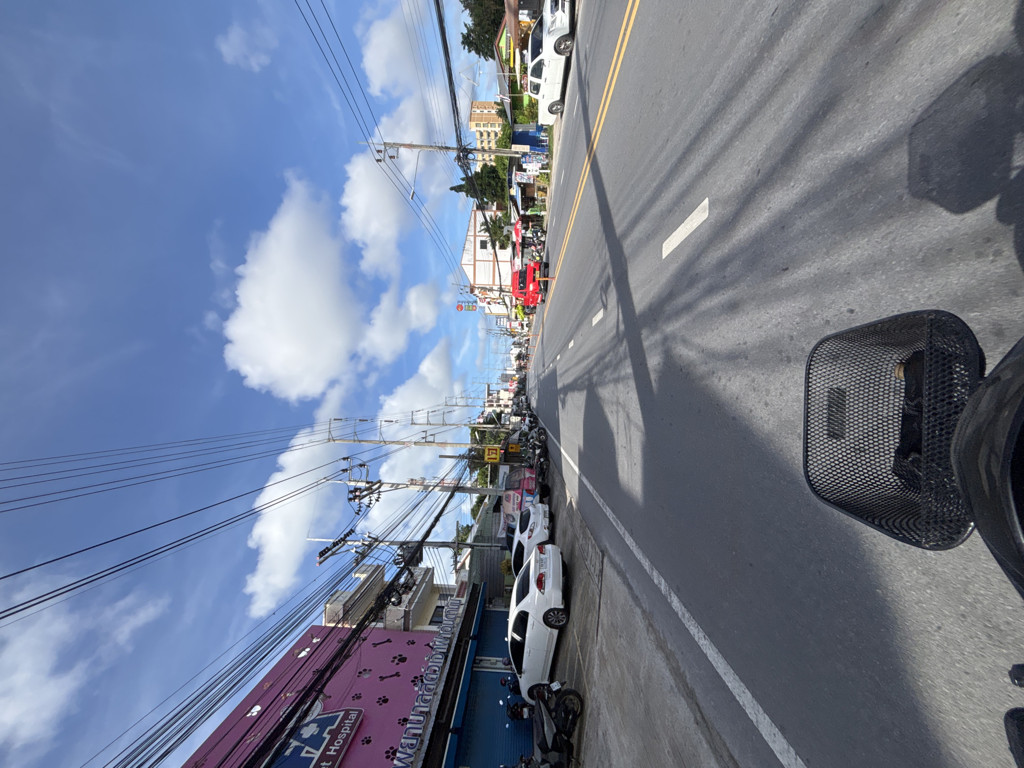 A first-person perspective shot, likely from a motorcycle or scooter, shows a street scene in Chalong, Thailand. The road is paved with asphalt and marked with double yellow lines and white dashed lines. On the left side of the road, a pink building with "Pet Hospital" written on it is visible, along with parked motorcycles and white cars. A row of shops and other buildings line the right side of the street. Overhead, numerous power lines crisscross the sky, casting shadows on the road. The sky is blue with scattered white clouds. A black basket is in the foreground, obscuring part of the view.