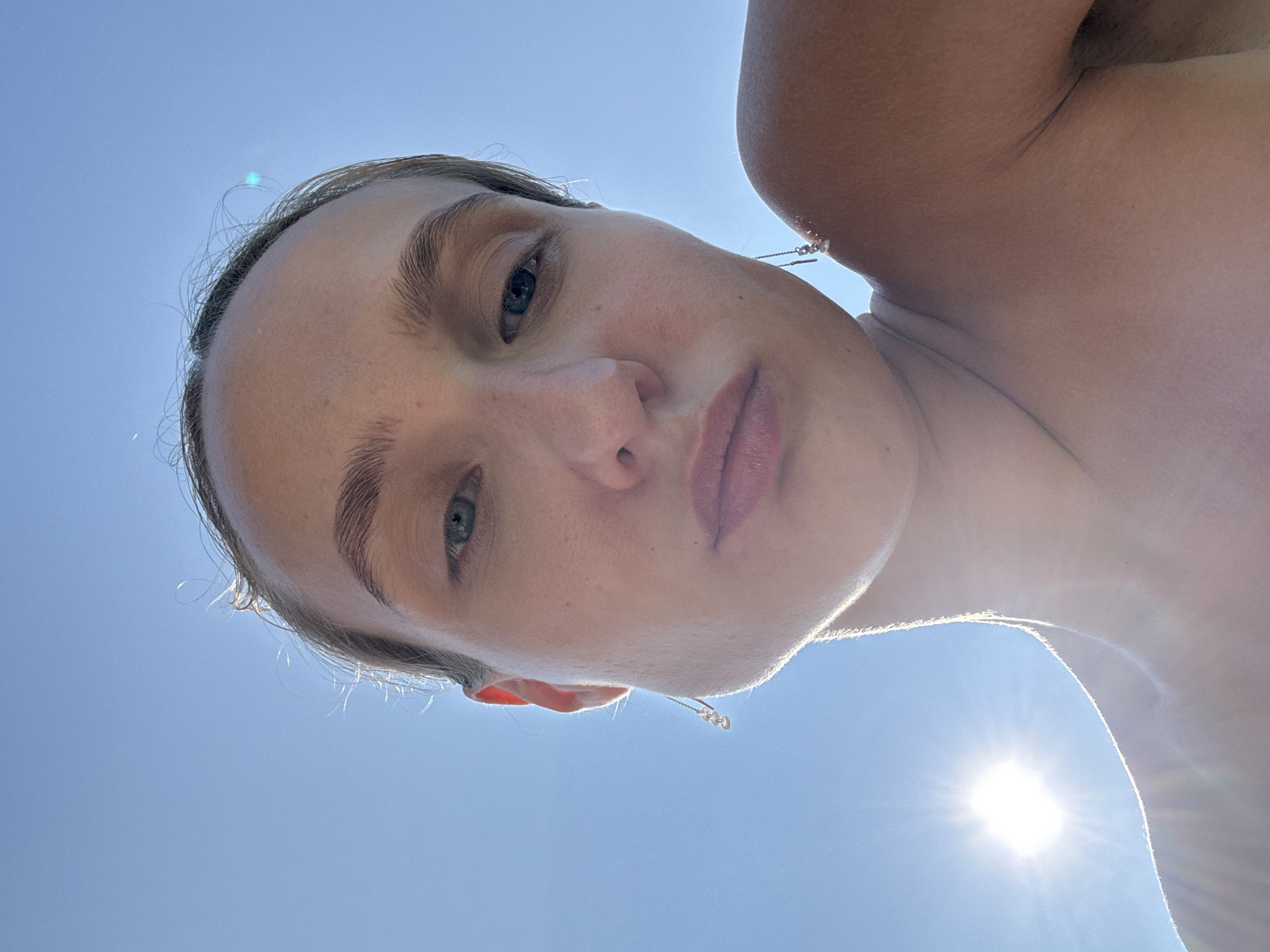 The image shows a close-up, low-angle selfie of a young woman against a bright blue sky. The sun is visible on the left side of the frame, creating a lens flare effect and strongly illuminating the subject's face and shoulder from the side and behind. The woman's face is the primary focus, with her eyes looking slightly upward and to the left. She has light brown hair pulled back, and is wearing dangling earrings. Her bare shoulder and upper chest are visible on the right side of the frame. The image appears to have been taken outdoors, possibly on a sunny day at a beach or other open area. There is no indication of the location being in the city of Подшивалово, Russia.