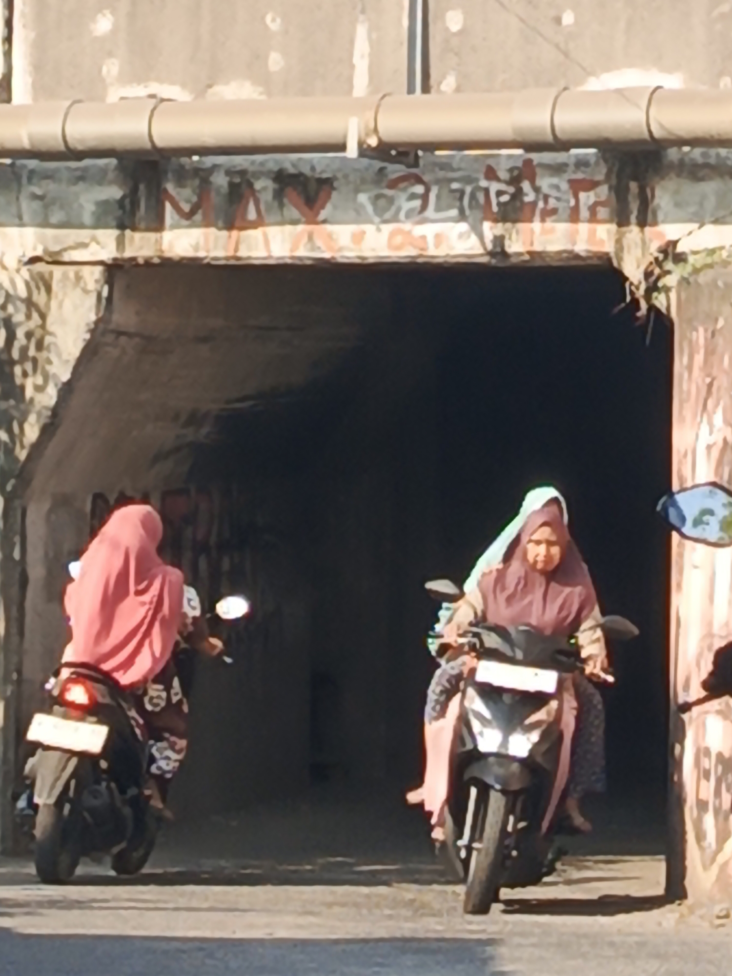 Two women wearing hijabs are riding motorcycles. The woman on the left is wearing a pink hijab and is riding a black motorcycle away from the camera, entering a tunnel. The woman on the right is wearing a light blue and maroon hijab and is riding a black motorcycle towards the camera, exiting the tunnel. The motorcycles are side-by-side. The tunnel appears to be under a concrete structure with a pipe running along its top. There is graffiti on the walls of the tunnel entrance and the exterior structure. The scene appears to be outdoors, with sunlight casting shadows. The location is identified as Kludan, Indonesia.