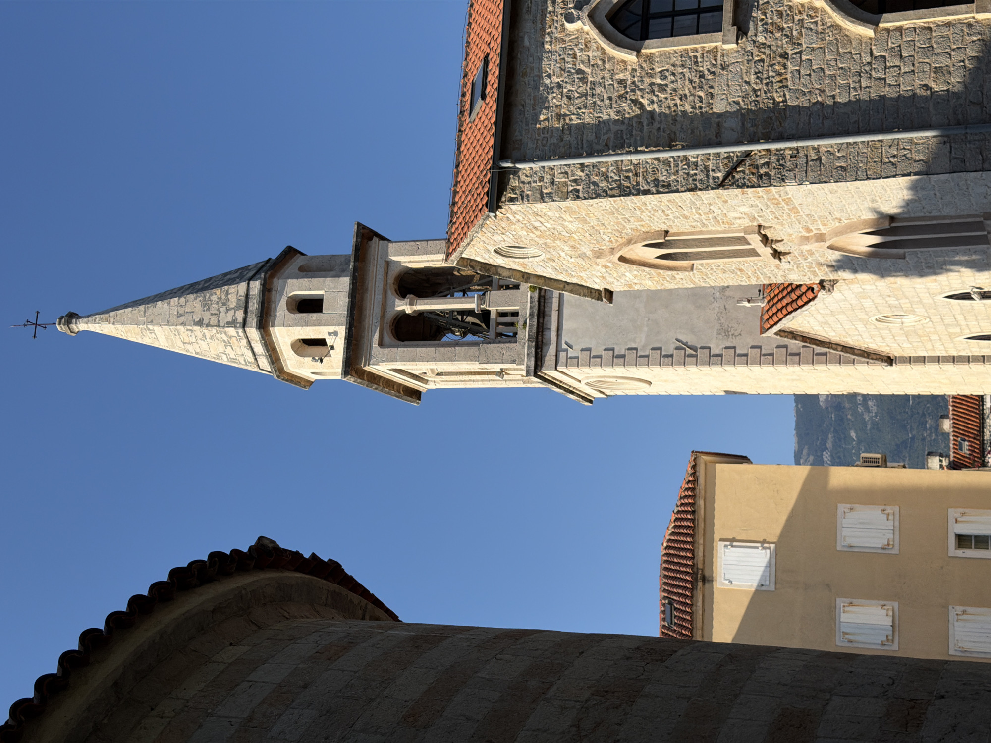 This is a photograph of the Church of Saint John in Budva, Montenegro. The image is taken from a low angle, looking up at the church tower and parts of its facade, framed by two other buildings. The sky is a clear, vibrant blue.
On the left, a curved building with a red tiled roof is partially visible. Its facade is made of stone with horizontal bands of a darker color.
In the center of the image is the bell tower of the Church of Saint John. It is a tall, slender structure constructed of light-colored stone. The tower has a pointed, spired roof topped with a cross. Below the spire, there are arched openings in the bell tower, revealing bells within. The tower is adorned with decorative stone elements and has multiple levels.
To the right of the tower is a section of the church building. This part of the church is also constructed of light-colored stone, with a rough, textured surface. It has a red tiled roof with a small dormer window. The facade features arched windows, some of which appear to be Gothic in style.
In the background, between the church and the building on the left, a light yellow building with white shutters is visible. It has a red tiled roof and appears to be several stories high. Beyond this, distant mountains can be seen.
The lighting suggests it is daytime, likely late afternoon, as indicated by the shadows cast on the buildings. There are no people visible in the frame. The scene depicts historic architecture bathed in sunlight against a clear blue sky.