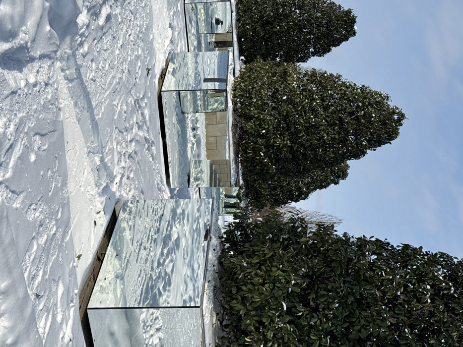 The image displays an outdoor winter scene in the city of Plodorodny, Russia, under a clear blue sky. The ground is extensively covered with white snow, exhibiting numerous footprints and disturbed areas. Several large, evergreen trees with dense, dark green foliage are situated within distinct, rectangular, cube-shaped planters. These planters are completely clad with highly reflective mirror surfaces. The mirrored surfaces reflect the snow-covered ground, the blue sky, and other planters and trees, creating fragmented visual effects. Three such mirrored planters are prominent in the foreground and midground, each containing a tree. The reflections also show segments of a light-colored, possibly beige, wall or structure in the background. A partial reflection of human legs, clad in blue jeans, is visible on one of the more distant mirrored planters. The scene is brightly illuminated by natural sunlight, casting subtle shadows.