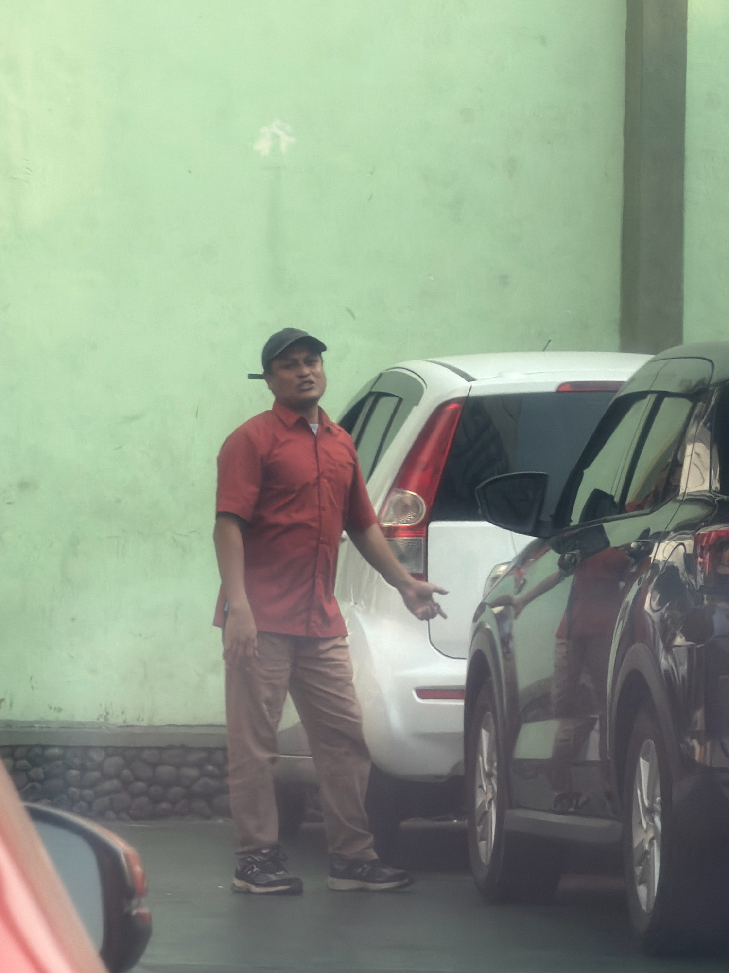 A man wearing a red collared shirt, brown pants, a black baseball cap, and dark sneakers stands to the left of a white car. He is facing towards the left of the frame and gesturing with his right hand. A dark car is parked next to the white car on the right. The scene appears to be outdoors with a pale green wall in the background.