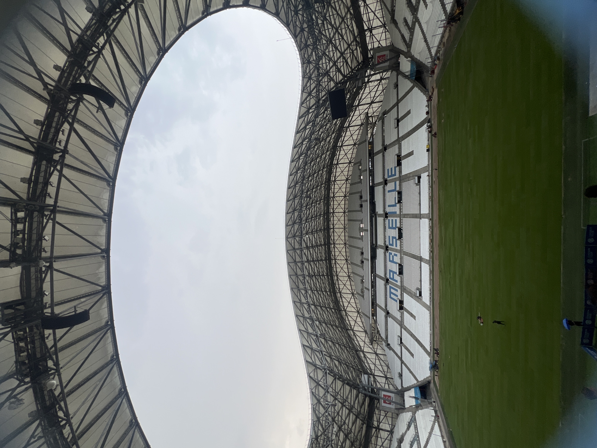 The image shows a view inside the Orange Vélodrome stadium in Marseille, France. The stadium features a large, undulating roof structure with a complex metal framework and a white translucent covering. White seating is arranged in tiered sections around a green football pitch. The word "MARSEILLE" is visible in large blue letters across the seats in the stands. The sky is overcast and appears pale grey. On the pitch, several individuals are present, with some appearing to be engaged in maintenance work, including the use of machinery. One person is standing in the middle of the pitch, and another is walking near the edge. In the foreground, a person holding a blue umbrella is visible near a barrier with signage. The overall scene suggests an empty or minimally populated stadium, possibly during an off-season or before an event.