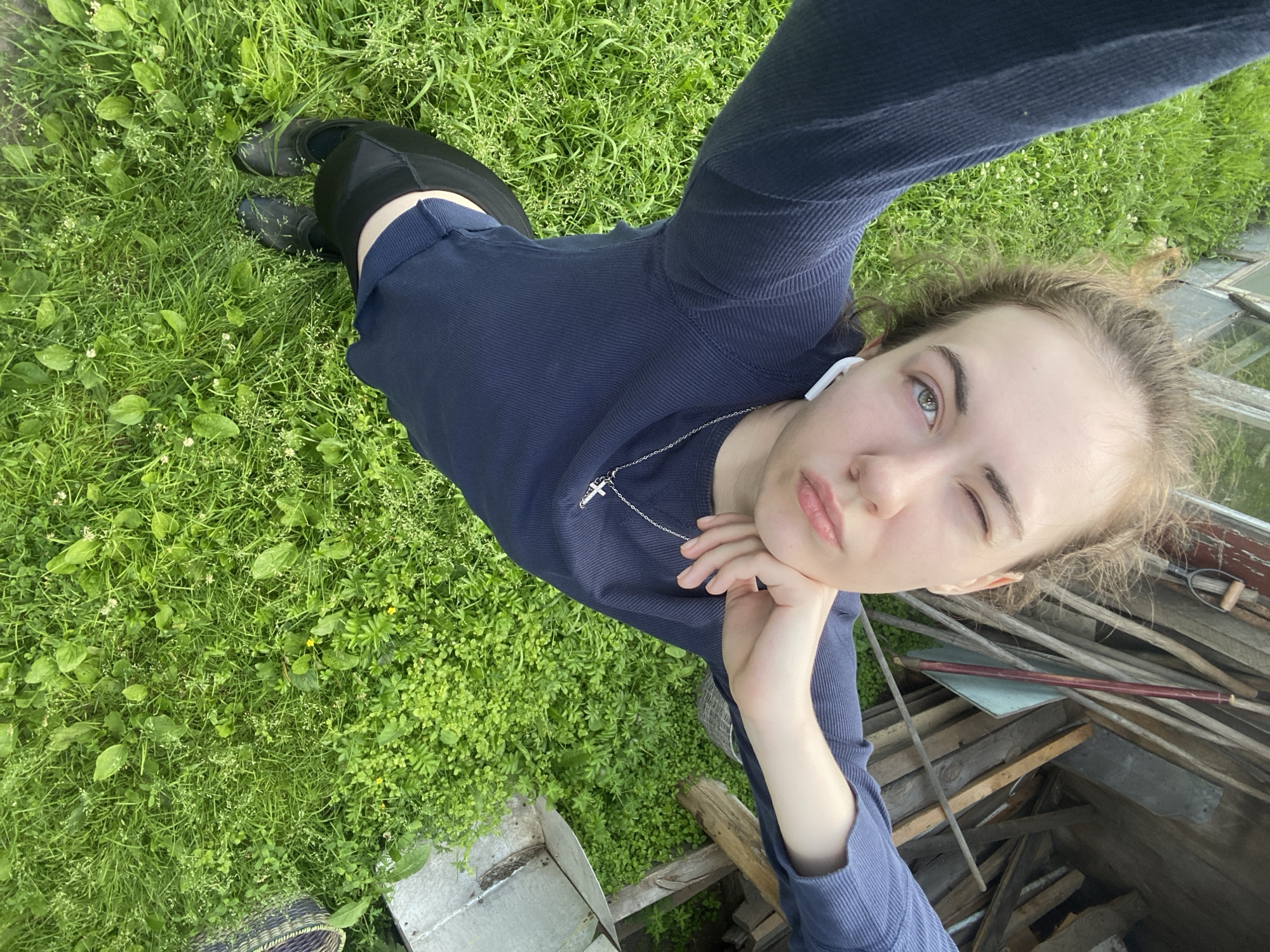 The image shows a young person lying on lush green grass, taking a selfie. The person is wearing a dark blue long-sleeved shirt and black pants, with black shoes. They have light-colored hair and are wearing white earbuds. A silver necklace with a cross pendant is visible around their neck. The person's right hand is resting on their chin, and they are winking at the camera. Their facial expression suggests a playful or casual mood.
The setting appears to be outdoors, with a focus on the natural environment of grass and plants. In the background, there are some wooden structures and what looks like a greenhouse, suggesting a rural or garden setting. The lighting indicates it is daytime, possibly overcast or late afternoon, as there are no harsh shadows.
The image is taken from a high-angle perspective, looking down at the subject. The overall impression is one of a relaxed and informal moment captured outdoors. No text is visible in the image.