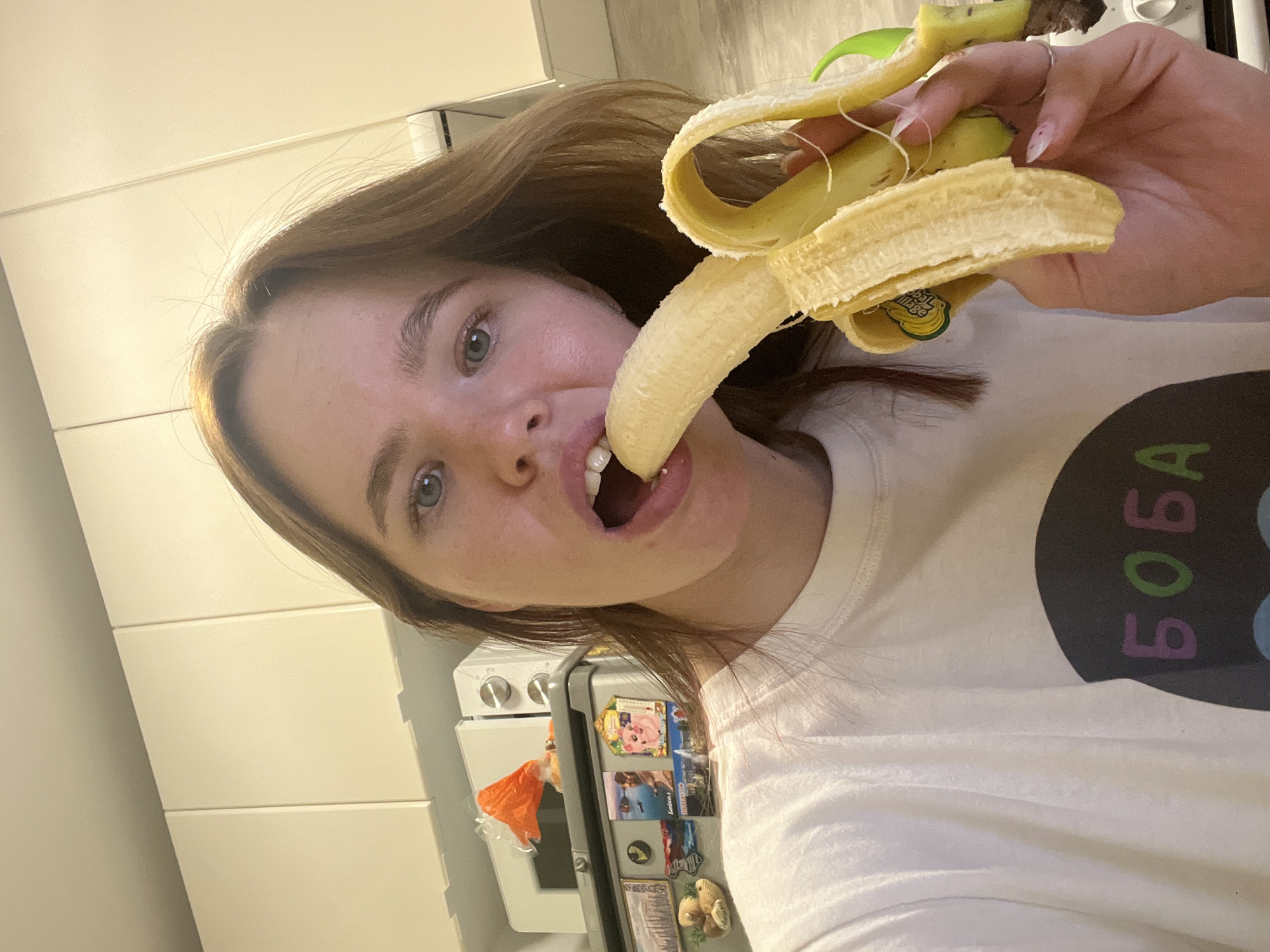 A fair-skinned person is holding and eating a peeled banana. The banana's peel is partially removed. They are wearing a white t-shirt with the text "BOBA" written vertically. The setting appears to be a kitchen, with white cabinets visible in the background. A refrigerator is behind the person with magnets affixed to it. The location in the city of Chaikovsky, Russia is unknown.