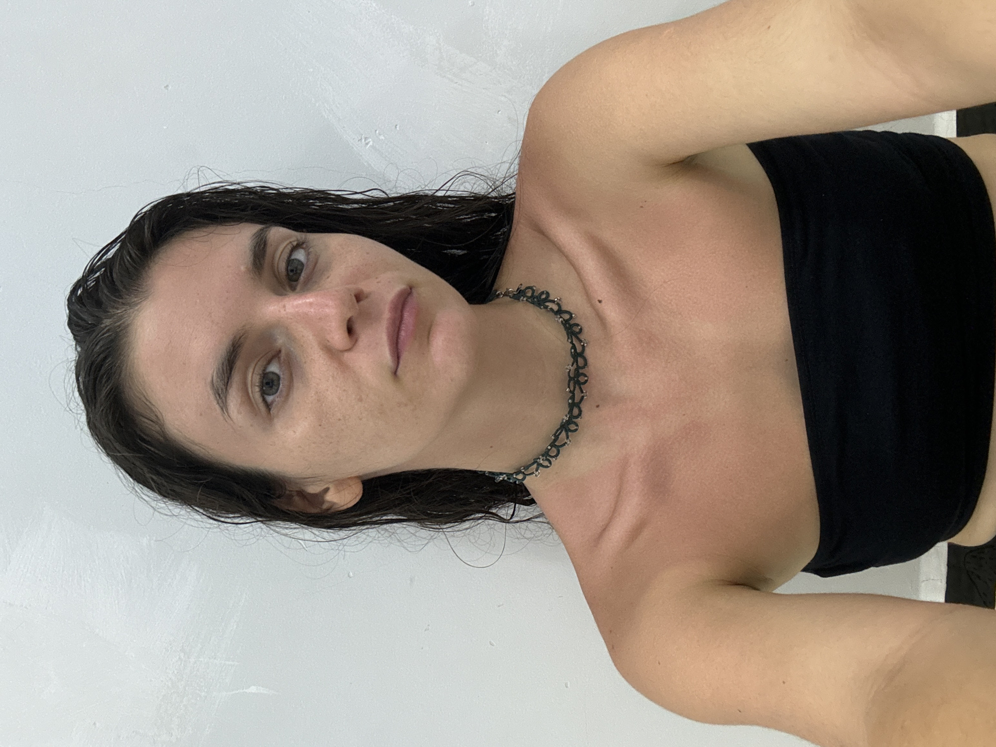 A medium-angle shot of a Caucasian woman with wet dark hair, facing the camera with a slight tilt. She is wearing a black strapless top and a dark choker necklace. Her skin appears slightly reddened. The background is a plain white wall. The location is not discernible from the image. There is no information available to suggest this photograph was taken in Mũi Né, Vietnam.