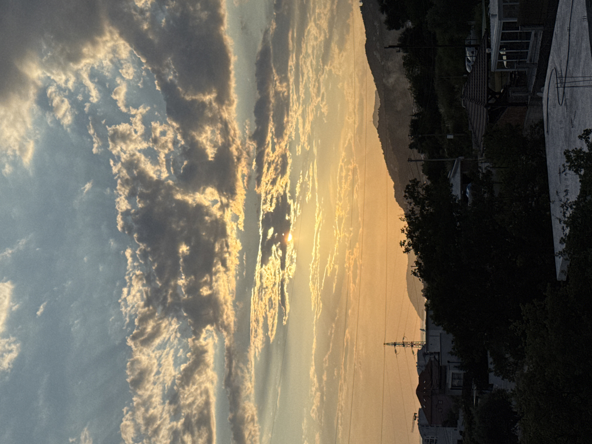 This is a vertical, eye-level, medium shot of a sunset over a suburban area in Sustaš, Montenegro. The sky dominates the upper two-thirds of the image, filled with dramatic clouds illuminated by the setting sun. Rays of golden light pierce through breaks in the clouds, creating a dramatic and vibrant display. Below the sky, a silhouetted landscape of rolling hills meets the horizon. In the middle ground, trees and buildings are visible. A tall electricity pylon stands prominently on the left side of the frame. The foreground consists of darker, silhouetted trees and rooftops, with one rooftop appearing to be under construction. The overall impression is one of a serene, yet active, evening scene in a coastal town.