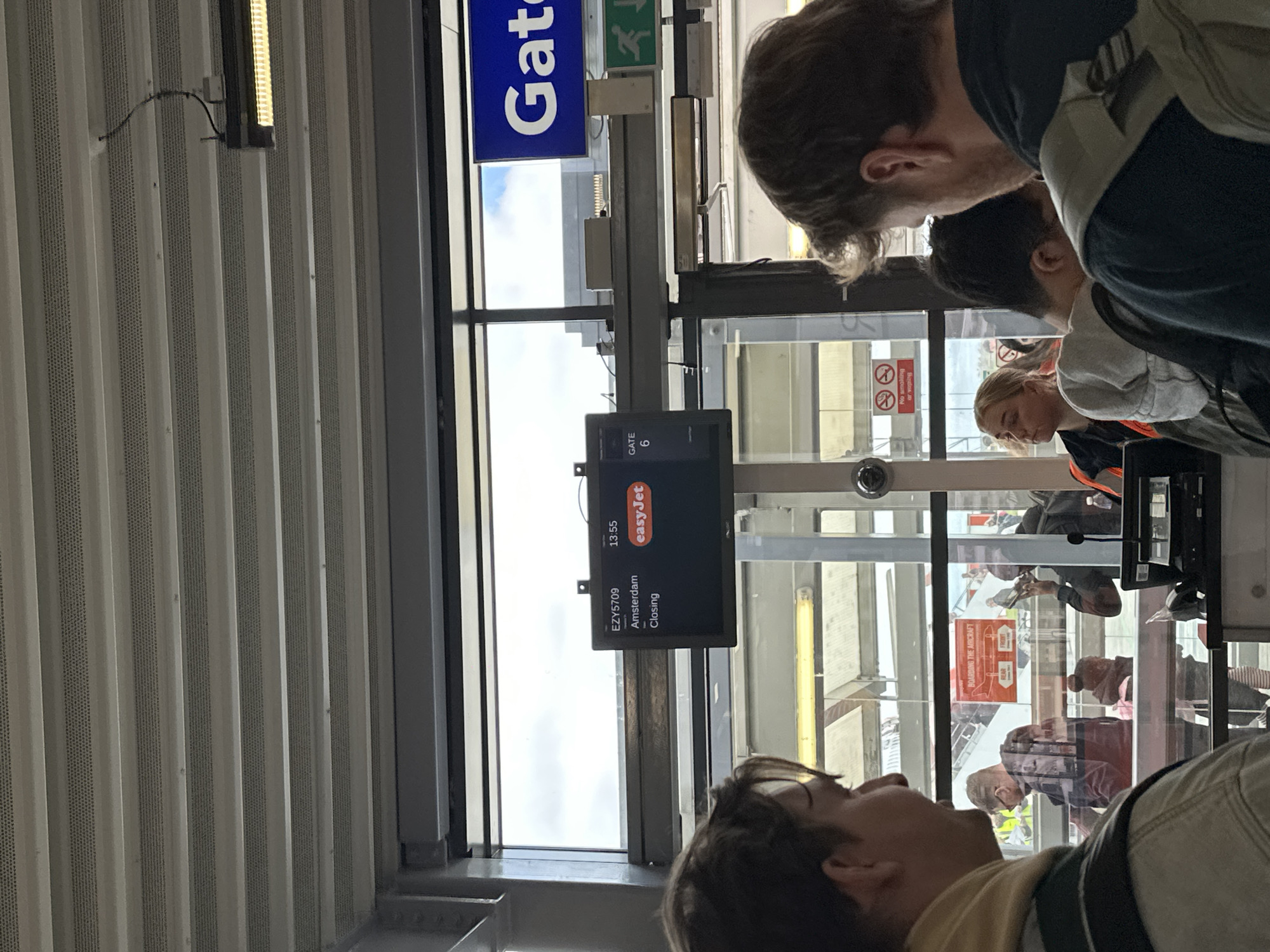 The image shows a scene at what appears to be an airport gate, potentially in Rochford, United Kingdom, judging by the presence of EasyJet branding. A digital display indicates that EasyJet flight EZY5709 to Amsterdam is closing and that it is 13:55. The sign also shows Gate 6. Several individuals are visible in the frame. The people are potentially passengers waiting to board or staff members. A window allows a view of the outside, and possibly other passengers. The environment is indoors, likely an airport terminal. The scene suggests a typical moment of travel and movement within an airport environment.