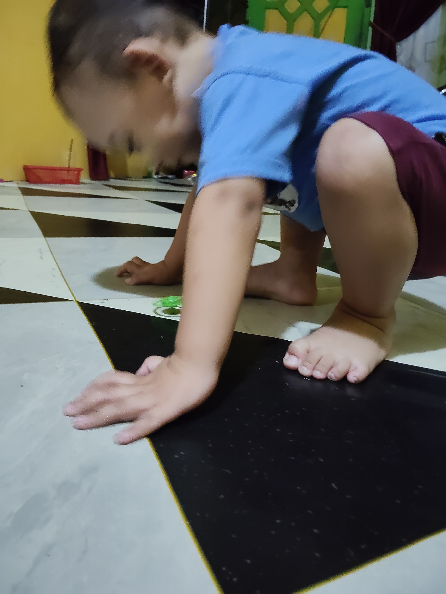 A young child, likely a toddler, is crouching on a checkered floor. The child is wearing a blue t-shirt and dark red shorts. Their body is angled towards the floor, with their hands placed in front of them for support. The floor has a pattern of black and white or light gray squares. There are hints of a yellow wall in the background, and a red plastic basket is visible further back. A green object is on the floor near the child's hands. The lighting suggests it is daytime indoors. The child's focus appears to be on something on the floor, but their face is not clearly visible. The overall scene depicts a child playing or exploring on the floor. The location context suggests this image was taken in Tanah Grogot, Indonesia.