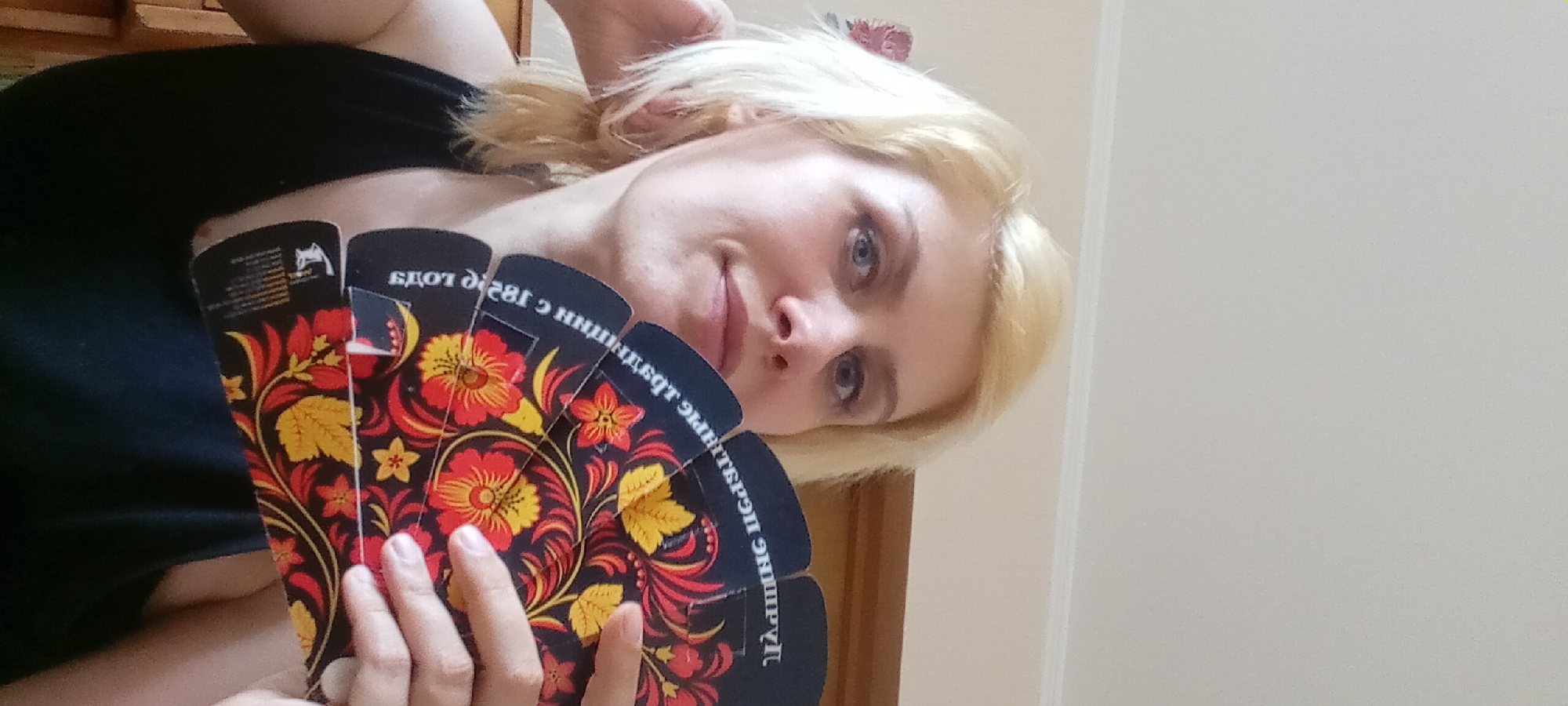 A blonde woman in a black tank top is holding a black fan with a red and yellow floral pattern. She is looking towards the camera. The background is a light beige wall.