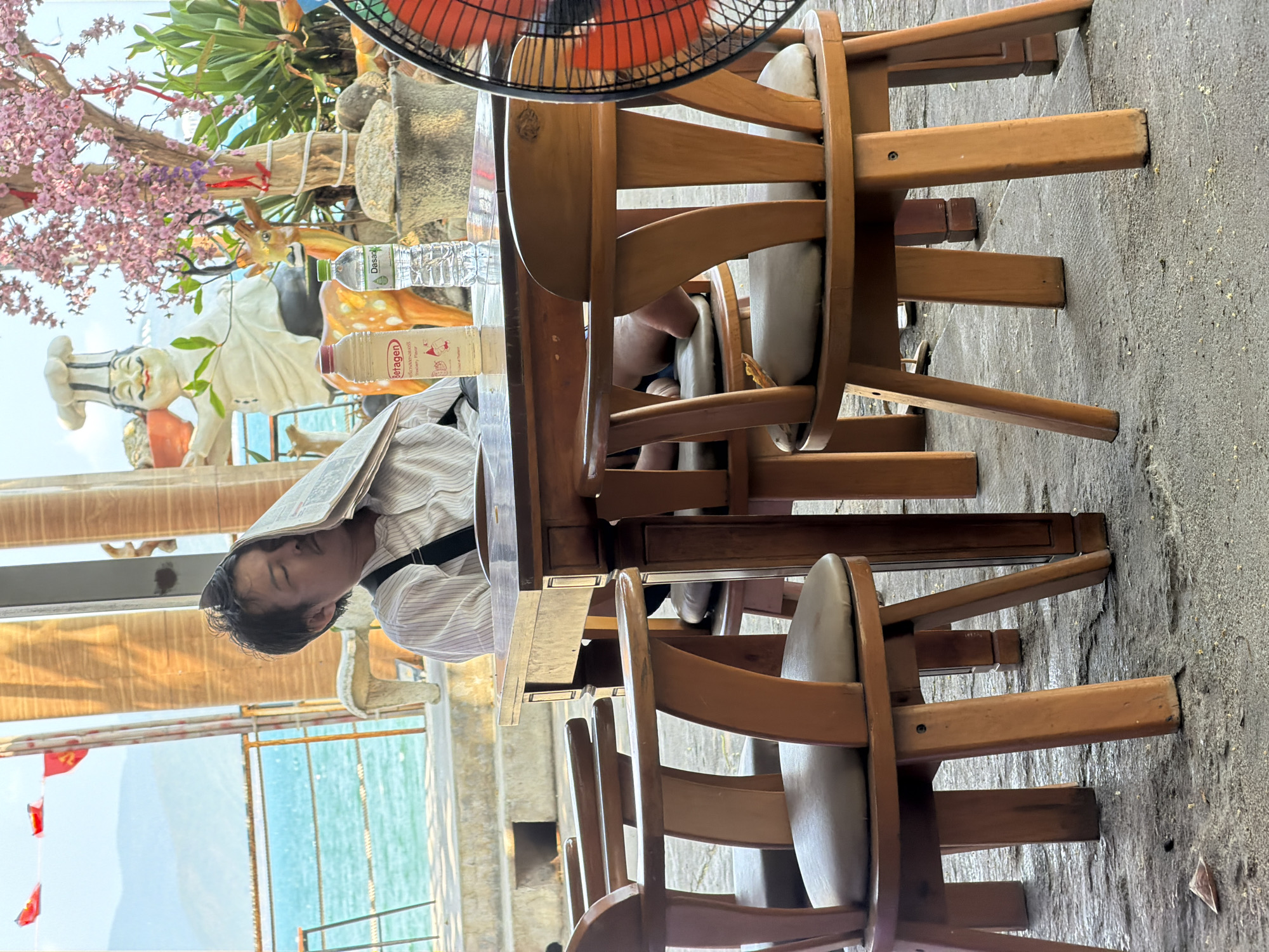 A man is sleeping at a wooden table in an outdoor setting with the edge of the ocean visible, likely in the Nha Trang Ward, Vietnam. The man has a newspaper covering his face, and he is wearing a striped shirt and suspenders. The table is set with bottled drinks and is surrounded by wooden chairs. A statue of a chef stands behind the table, along with potted plants and artificial flowers. A red electric fan is partially visible above the table. The ground is concrete.