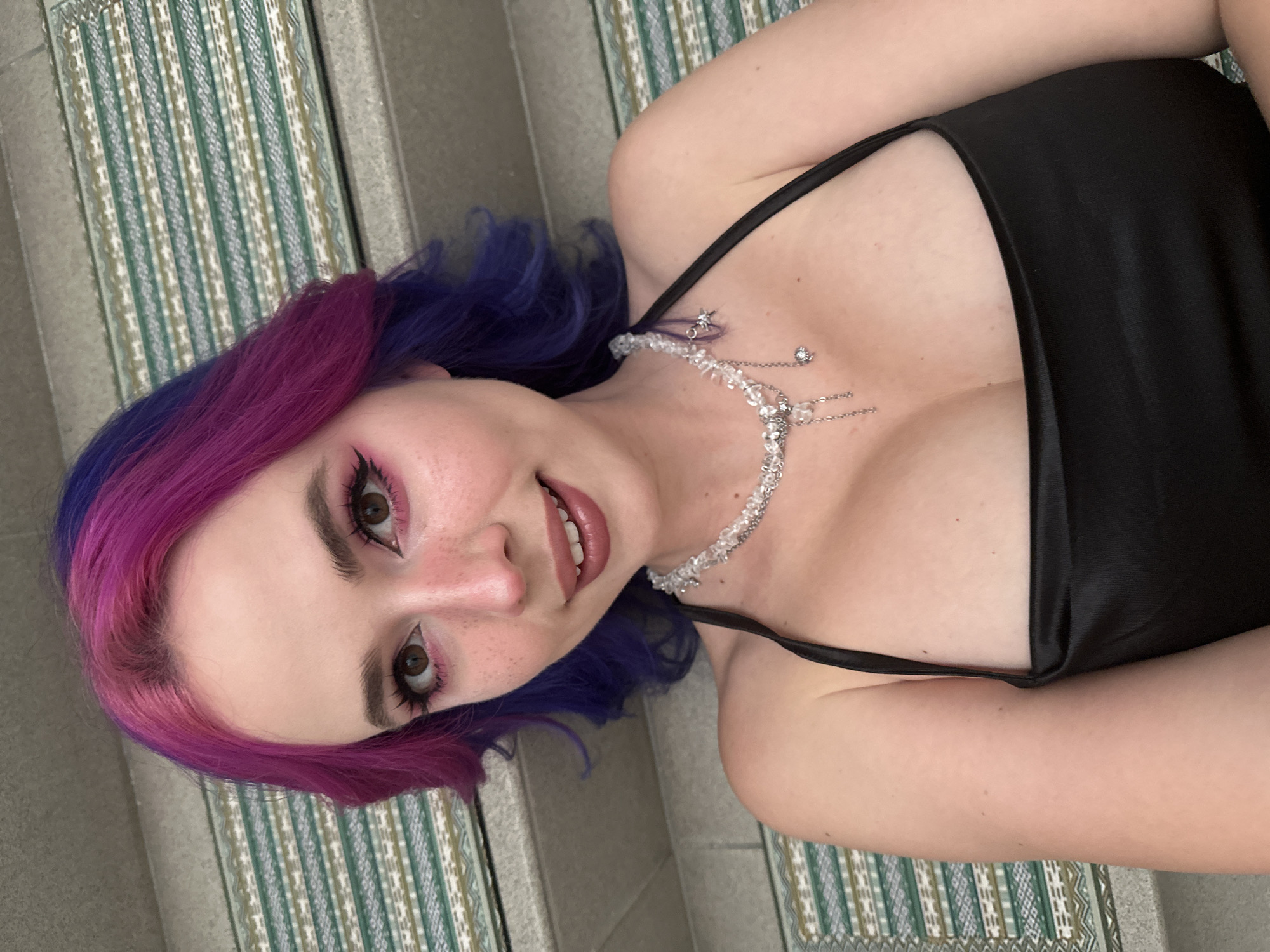 A close-up, tilted portrait features a young woman lying down, looking directly at the camera with a gentle smile. She has distinctive bi-colored hair, transitioning from dark purple to bright magenta/pink, styled in soft waves. Her fair skin shows visible freckles across her nose and cheeks. Her brown eyes are enhanced with dramatic pink eyeshadow, dark eyeliner, and mascara. Her lips are a light, glossy pink or nude.
She is wearing a black, possibly satin, spaghetti-strap top or dress. Adorning her neck and décolletage is an intricate necklace composed of clear crystal shards strung together, connected by delicate silver chains featuring small star-shaped charms. One of the chains appears to extend towards an ear, possibly connecting to a piercing.
The background consists of a patterned surface, resembling a rug or mat, displaying vertical stripes and geometric designs in shades of green, white, and brown. This patterned area is adjacent to a plain gray surface, suggesting an indoor setting. The photograph is taken from a slightly elevated and tilted perspective. No visible text is present in the image. The location context for this scene is the City of Zagreb, Croatia.