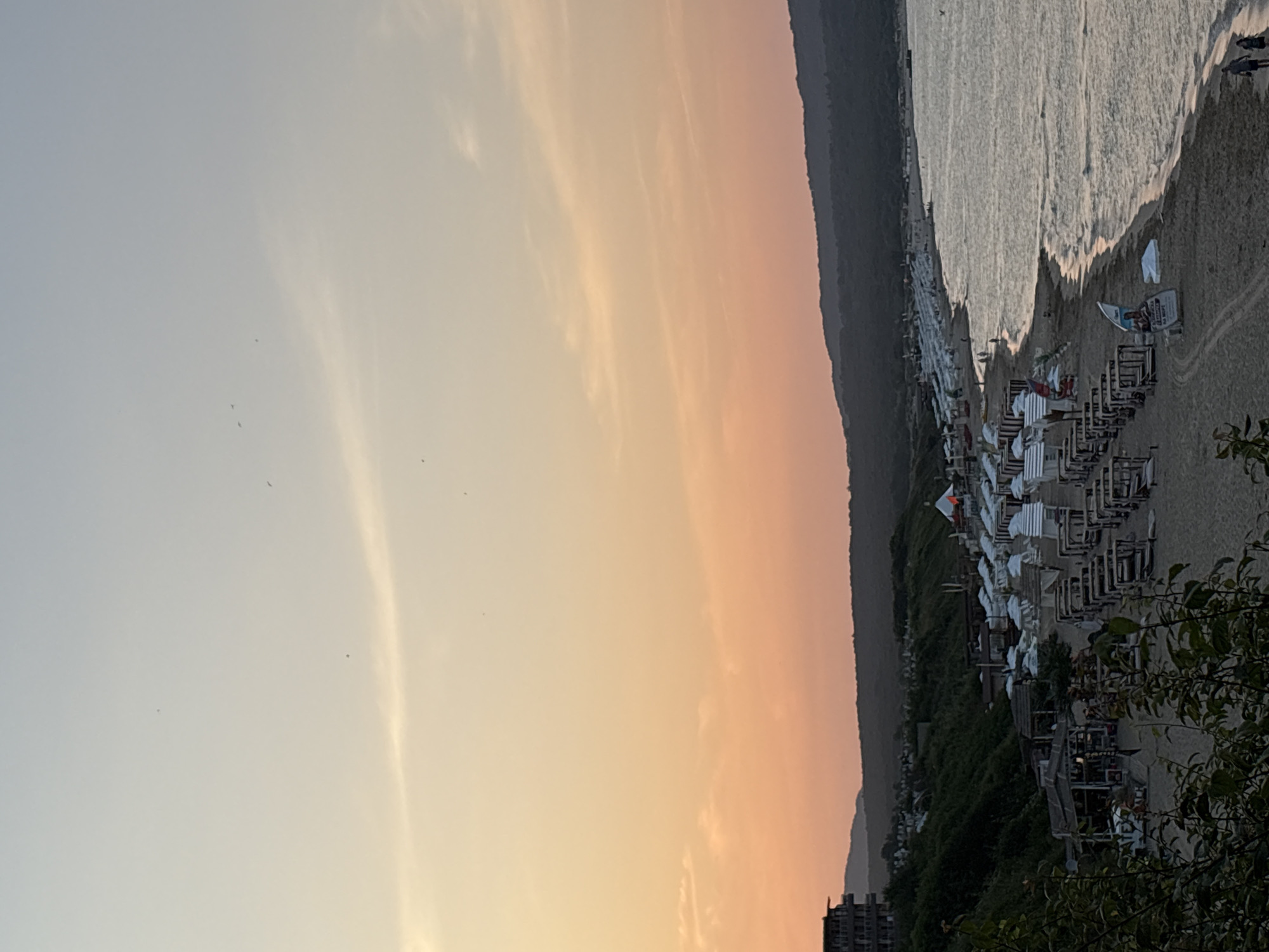 The image depicts a wide, sandy beach on the coast of Primorsko, Bulgaria, during a sunset or sunrise. The sky displays a gradient of soft orange and pink hues, with a few wispy clouds. The sea is calm with gentle waves reflecting the sky's colors.
The beach is lined with numerous rows of white umbrellas and lounge chairs, indicating a popular tourist destination. Some of these appear to be in cabana-style setups with wooden frames. There are also several small wooden structures on the beach, possibly kiosks or small bars. In the background, a dense green hillside rises, dotted with trees. A large, multi-story building, likely a hotel or resort, is visible on the left side of the image, situated on the hillside overlooking the beach.
A few people are present on the beach and near the water's edge. Two individuals are walking on the sand in the lower right corner, and others can be seen dotted along the shoreline and near the beach setups. The overall scene suggests a peaceful, serene atmosphere at the end of a day or the beginning of a new one at this Bulgarian coastal resort.
