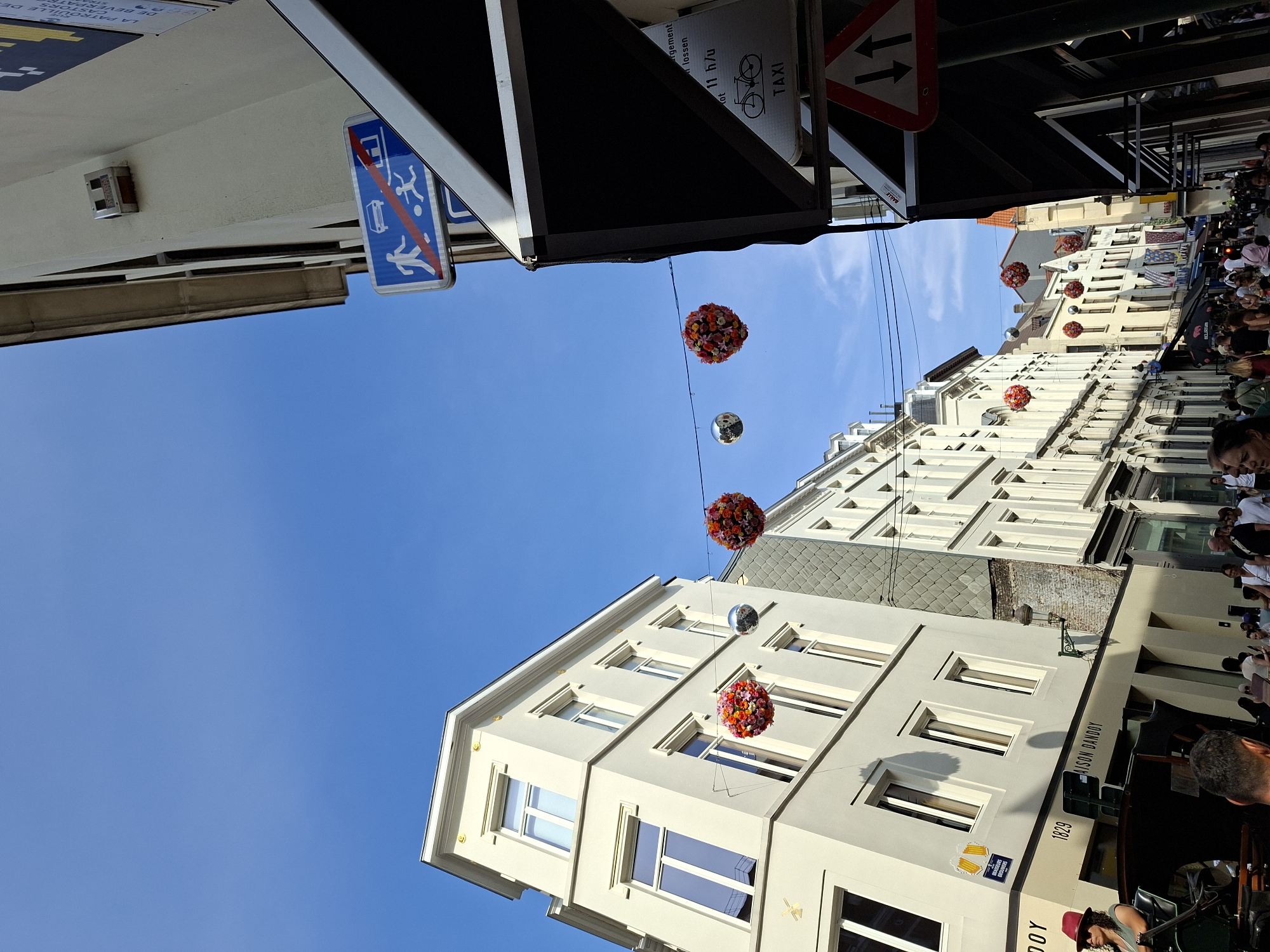 The image captures a bustling street scene in Brussels, Belgium, on a clear, sunny day. The perspective is looking up along a narrow street lined with tall, light-colored buildings. Decorative elements, specifically brightly colored floral spheres and a disco ball, are suspended by wires across the street, adding a festive atmosphere.
On the left, a prominent light-colored building displays architectural details like a stepped cornice and ornate window surrounds. A sign on this building reads "MAISON DANDOY" and "1829", indicating a business with a long history, likely a biscuit maker given the historical reputation of Maison Dandoy. Below this, a horse-drawn carriage is visible, with a woman wearing a red hat seated in it. Several people are gathered on the sidewalk near the carriage and the entrance to Maison Dandoy.
The right side of the street features more buildings, including an awning that partially obscures the view. Various signs are affixed to the buildings, including a blue traffic sign indicating a pedestrian and cyclist zone with a car symbol crossed out, suggesting it may be a restricted area for vehicles. Another sign displays a bicycle symbol and the word "TAXI". Further down the street, more people are walking and browsing shops, suggesting it is a commercial area. The sky is a vivid blue with a few wisps of clouds. The overall impression is of a lively urban environment with elements of both historical charm and modern commerce.