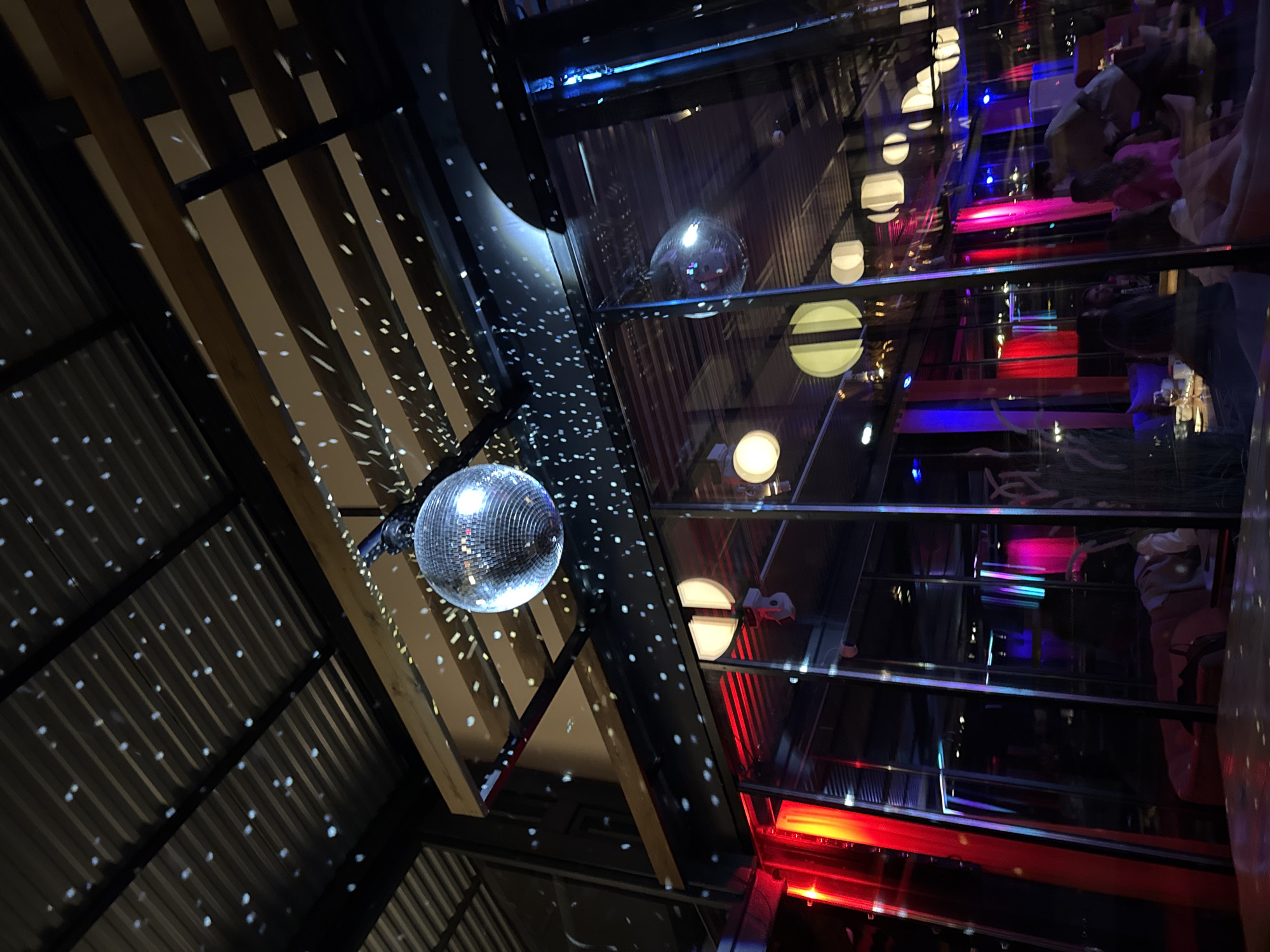 The image displays a vibrant indoor scene, likely a bar, club, or restaurant, characterized by a dark and dynamic atmosphere. A large disco ball hangs prominently in the upper-left quadrant, reflecting numerous small, bright light spots across the corrugated metal ceiling and exposed wooden beams.
On the right side of the frame, large glass panels or windows reflect and reveal parts of the interior. Through and in these glass surfaces, multiple round, illuminated light fixtures are visible, along with distinct streaks of colored light in red, blue, and pink/purple tones, contributing to a lively ambiance. Reflections of indistinct figures, presumably people, can be seen in the glass, suggesting a social gathering or event. Some reflected individuals appear to be wearing colorful clothing, such as pink. The overall lighting is dim, indicating an evening setting or an interior reliant solely on artificial illumination. The architectural style blends industrial elements like the corrugated ceiling with natural wooden beams and modern glass partitions. No discernible text is visible in the image. The scene evokes a festive and energetic mood.