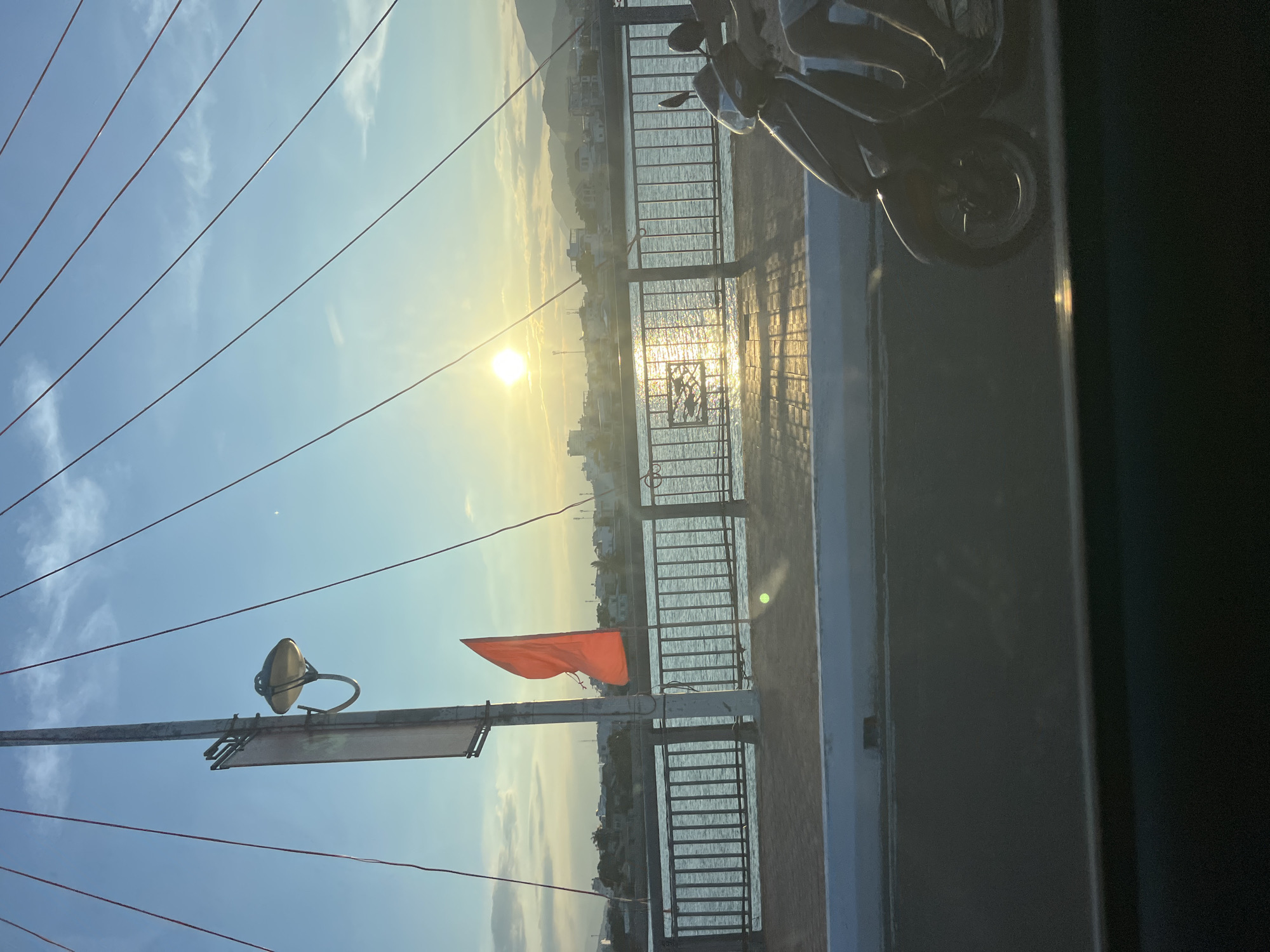 The image shows a scene on a bridge in Nha Trang Ward, Vietnam. The sun is low, reflecting on the water and illuminating buildings in the distance. A red Vietnamese flag is prominent on a pole to the left. Power lines stretch across the sky. A motorcycle is parked partially in the frame on the right side of the image, parked by the railing.
