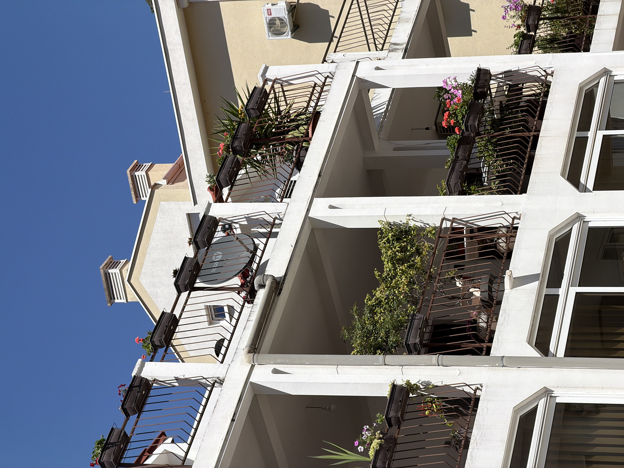 The image depicts a multi-story building in Budva, Montenegro, with a clear blue sky in the background. The building is constructed with white columns and balconies featuring brown wrought-iron railings. Each balcony is adorned with flower boxes containing various plants and flowers. A satellite dish is visible on one of the balconies, and an air conditioning unit is mounted on the exterior wall of the building. The overall aesthetic is modern and residential.