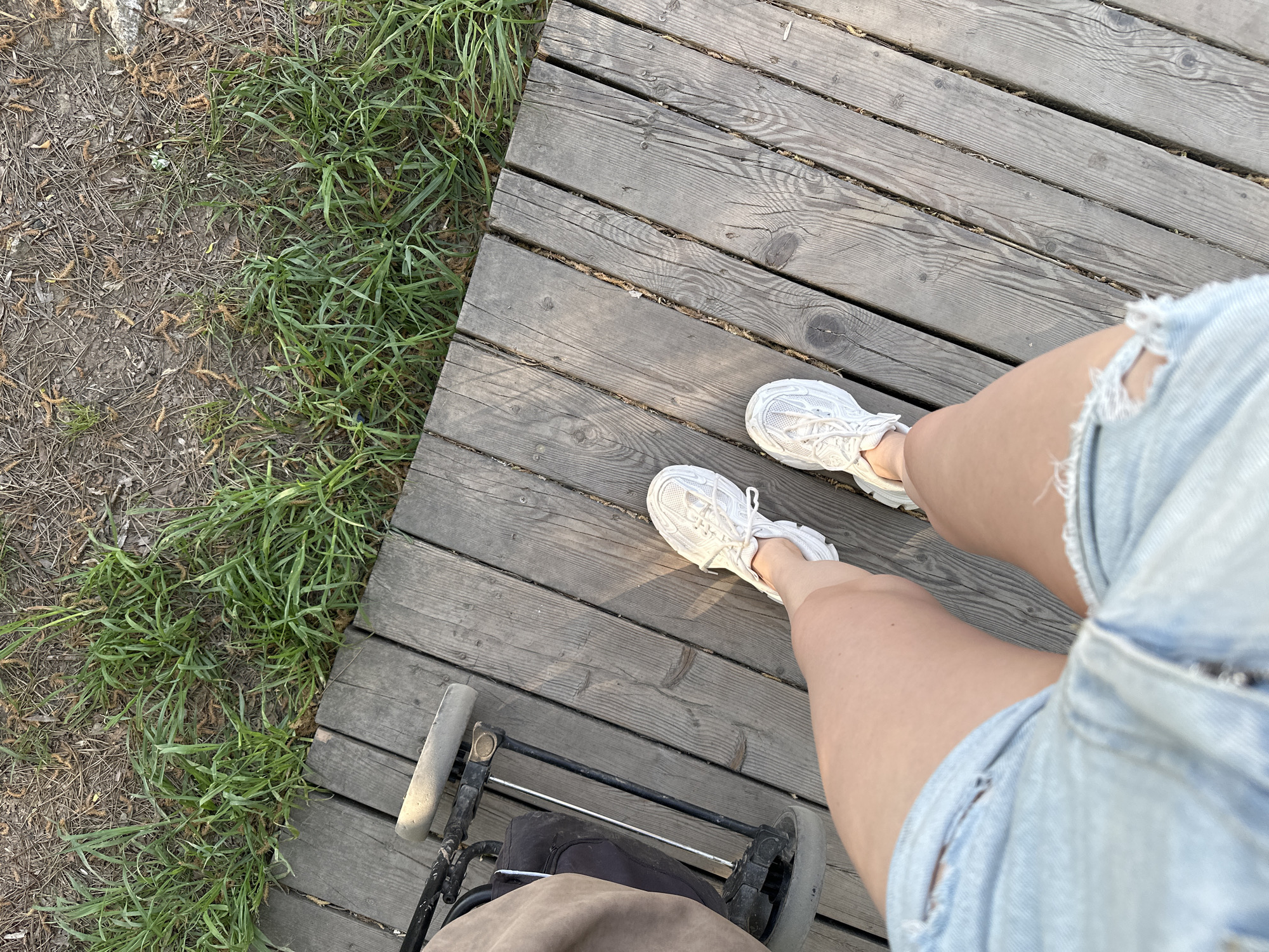 The image shows a person standing on a wooden boardwalk, next to a patch of grass and dirt. The person is wearing white sneakers and denim shorts. A stroller is in front of them. The specific location in the city of Kazan, Russia cannot be determined from the visual information.