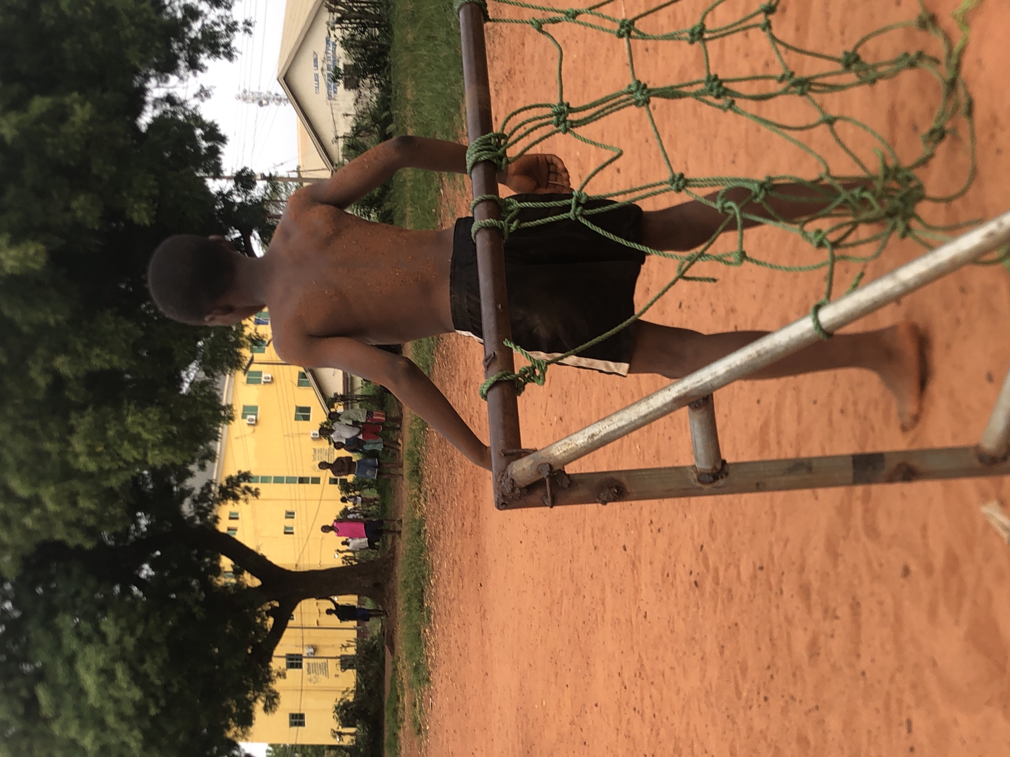 An outdoor, daytime image shows a young, shirtless person from behind, holding onto a green rope net attached to a rusty metal frame. The individual wears black shorts with white trim and stands barefoot on reddish-brown dirt ground, their skin appearing somewhat dusty.
In the mid-ground, a large, leafy green tree partially obscures a vibrant yellow, two-story building with numerous windows and green air conditioning units. Several people are gathered in front of this building, dressed in casual clothes, suggesting a social or observational activity. Further in the background, another lighter-colored building with a sloped roof is visible through the trees, above which faint power lines crisscross the sky.
Text on the lighter background building is partially legible and appears to include "COLLEGE LIBRARY" or "COLLEGE BUILD...", indicating an institutional or educational setting. The scene portrays a casual, communal atmosphere, consistent with a schoolyard or recreational area in Nsugbe, Nigeria. The activity of the foreground subject suggests play or interaction with a sports structure, possibly a goal. The time of day is daylight, under natural, diffused light.