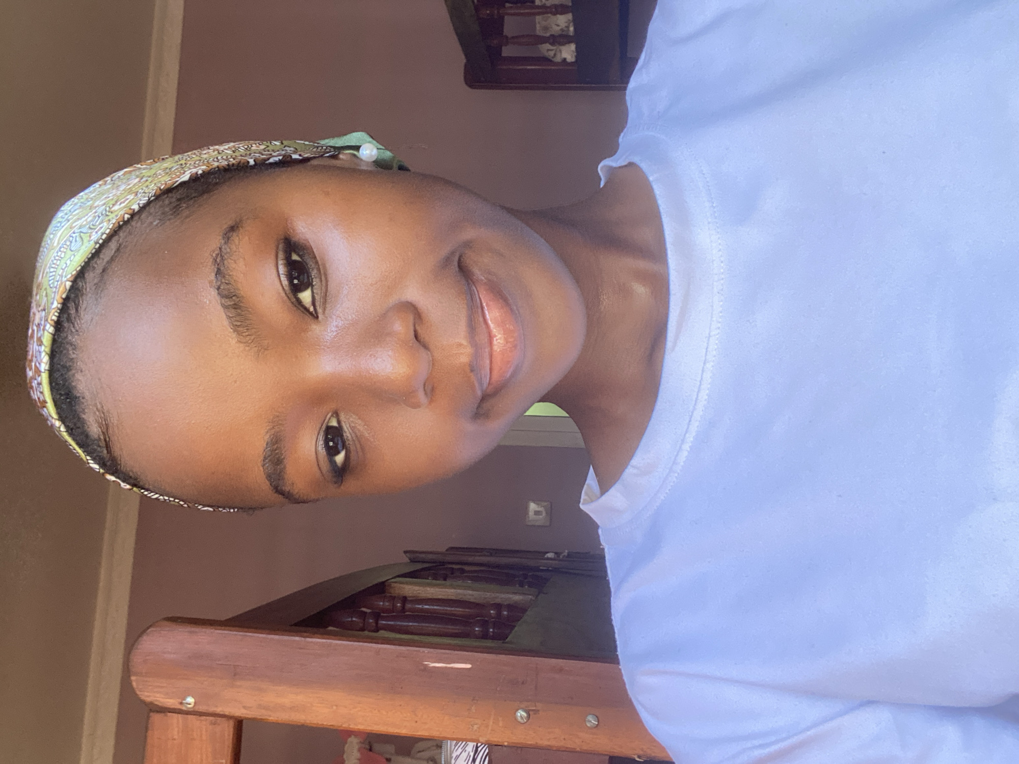 A medium-shot portrait of a young Black woman indoors. She is wearing a white t-shirt and a patterned headscarf. She has a neutral expression and is looking directly at the camera. The background is a dark color, and there is a window with natural light coming in. In the lower half of the image, the top of what appears to be an antique table or desk is visible. There is no evidence to indicate the location is Dar es-Salaam, Tanzania.