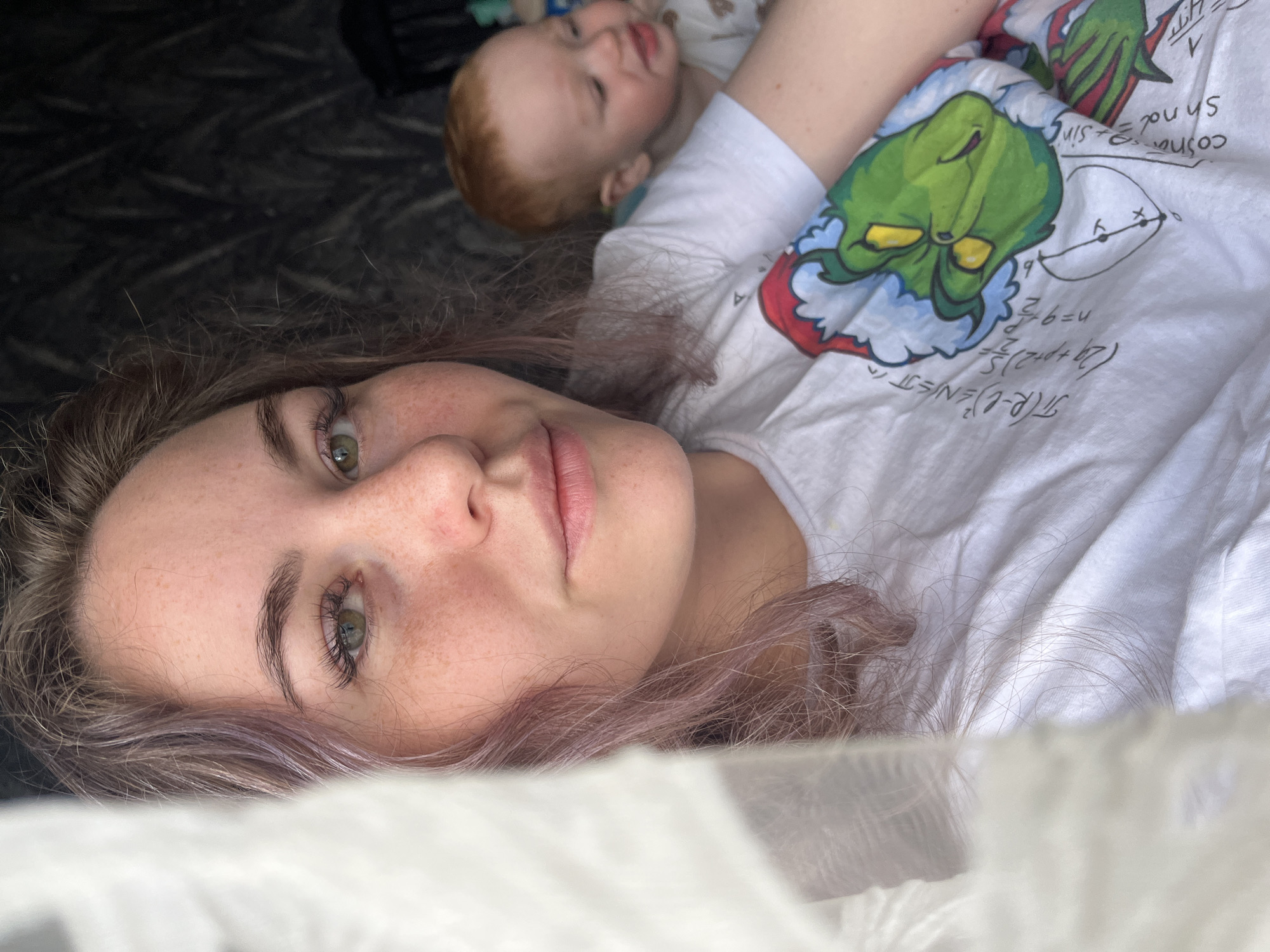 The image captures a relaxed indoor moment, likely in Chaikovsky, Russia, featuring a woman and a baby. In the foreground, a woman lies on her side, her head turned towards the camera. She has striking green eyes, subtle freckles across her nose and cheeks, and her hair is a distinct light purple or lilac shade, mixed with brown. Her expression is calm and neutral, with a slight hint of a smile.
Behind her, partially visible, is a baby with noticeable red or ginger hair, lying on its back. The woman is wearing a white t-shirt adorned with a festive graphic of the Grinch character, complete with a Santa hat, integrated into a design filled with mathematical formulas and a geometric diagram. Clearly visible text on the shirt includes: "(2g+p+2)= n=9 cosna Shnd= sin A". The background is dark and indistinct, while a light, possibly white or sheer, fabric occupies the immediate foreground, creating a soft bokeh effect. The lighting suggests it is daytime, contributing to the peaceful atmosphere of the scene. The image portrays a quiet, intimate moment between the two subjects.