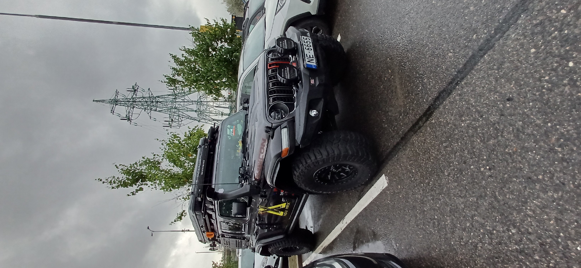 A black Jeep Wrangler is parked in a wet parking lot. The Jeep has a rooftop tent, a snorkel, and custom decals on the side. The license plate reads "NE-6655". In the background, there is a large electrical pylon and trees. The sky is overcast.