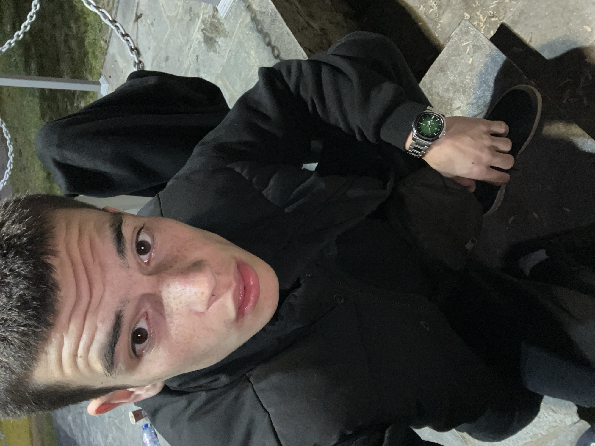 A young man is sitting on a gray concrete step in what appears to be an outdoor location. He is wearing a black hooded jacket and dark-colored pants. He has short dark hair and is looking upwards towards the camera. He is wearing a silver watch with a green face on his left wrist. His hands are clasped together. To his left, a chain railing is visible. A dark shoe is visible near his right hand. A water bottle is located on the ground in the lower left corner of the frame. The location is assumed to be Karavelovo, Bulgaria.