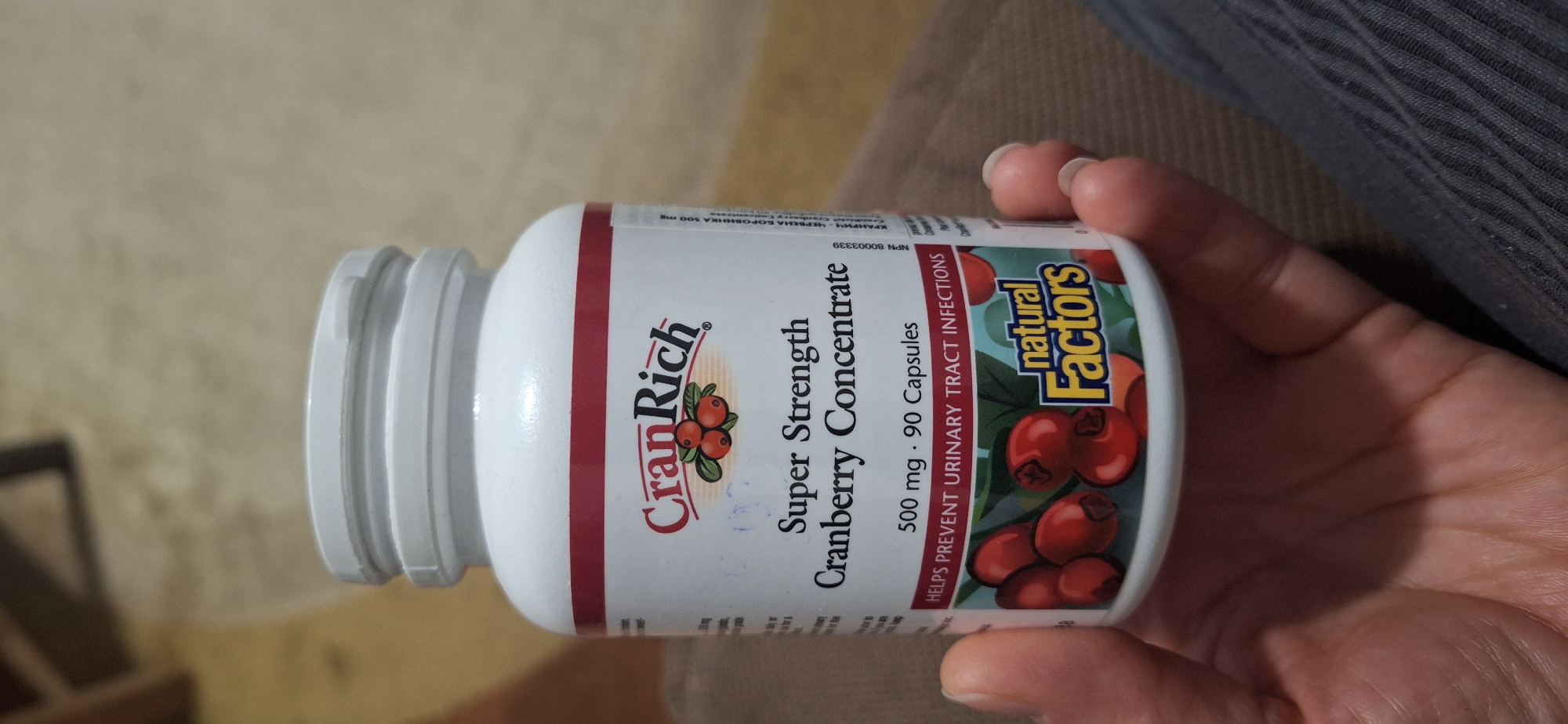 A close-up shot shows a bottle of "CranRich Super Strength Cranberry Concentrate" being held in a person's hand. The bottle is white with a red and tan label. The label prominently features the brand name "CranRich" in red lettering above an image of cranberries. Below, it reads "Super Strength Cranberry Concentrate" in black. Further down, it specifies "500 mg. 90 Capsules" and "HELPS PREVENT URINARY TRACT INFECTIONS." The right side of the label also has the "natural Factors" logo with an illustration of cranberries. The person's hand is visible, with pale skin and manicured nails. The background appears to be a neutral-colored fabric surface. There is no information in the image to suggest this is in Blagoevgrad, Bulgaria.