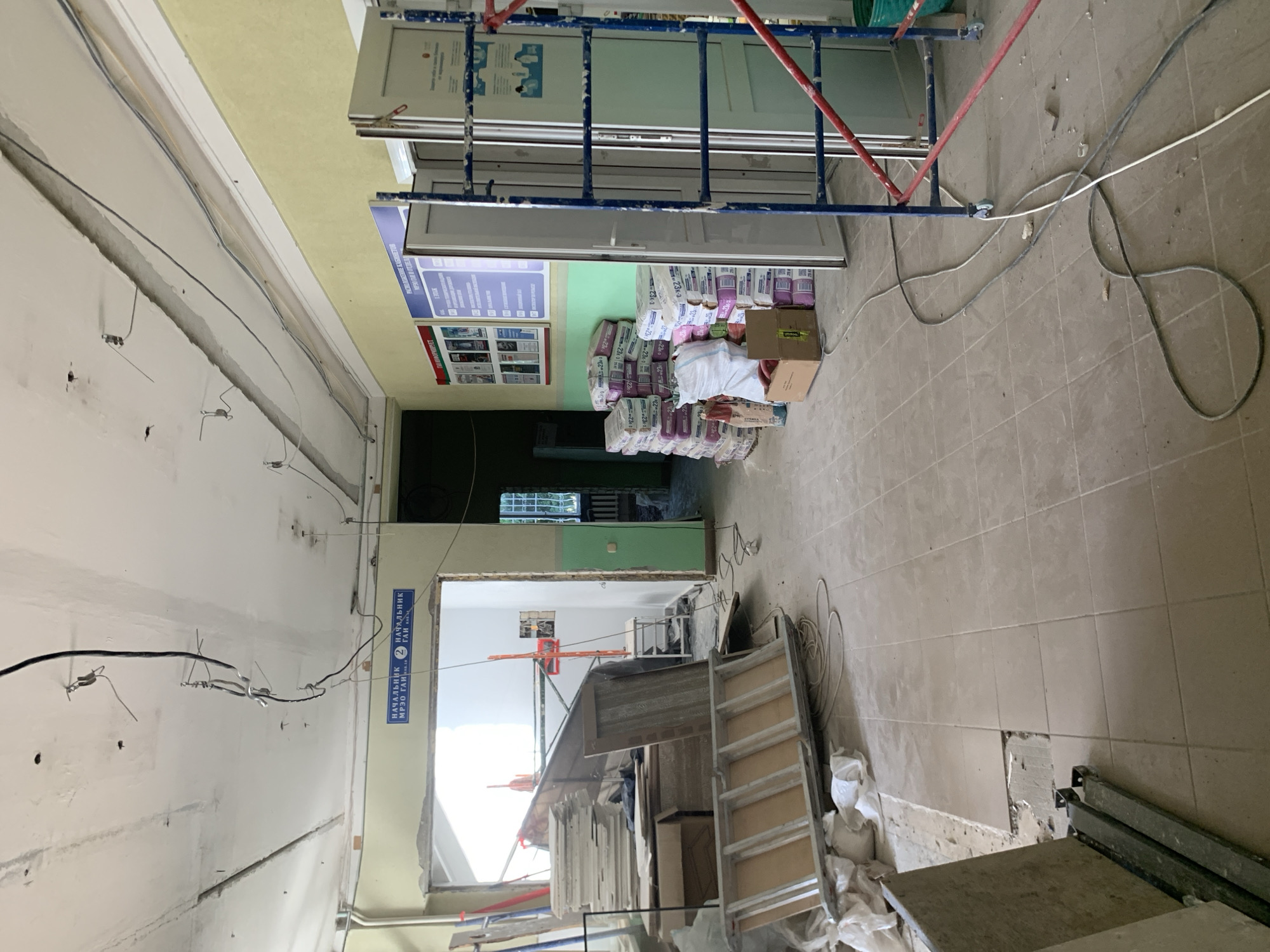 The image depicts an interior scene undergoing renovation or construction. The room appears to be a hallway or a large room with an unfinished ceiling where electrical wiring is exposed. The walls are painted in light yellow and green. On the left side of the image, there is a large opening that leads to another area, and construction materials such as stacks of tiles and wooden planks are visible. In the center and to the right, several stacks of bagged dry building mix are arranged against the wall, and a blue and red metal scaffolding is positioned in the foreground. There are also various cables and wires running across the tiled floor. A closed double door is visible on the right side of the image. The overall environment suggests an active construction site.