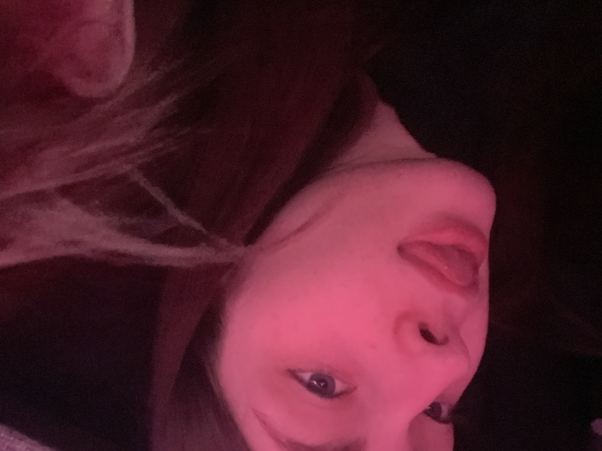 A close-up, slightly blurry image shows a person's face illuminated by pink lighting. The person's head is tilted, with their nose and mouth prominently displayed. The person's tongue is sticking out. One eye is open and visible, while the other is partially obscured. Parts of the person's hair are visible, along with dark surroundings. There are no discernible details that suggest a location within the city of Saint Petersburg, Russia.