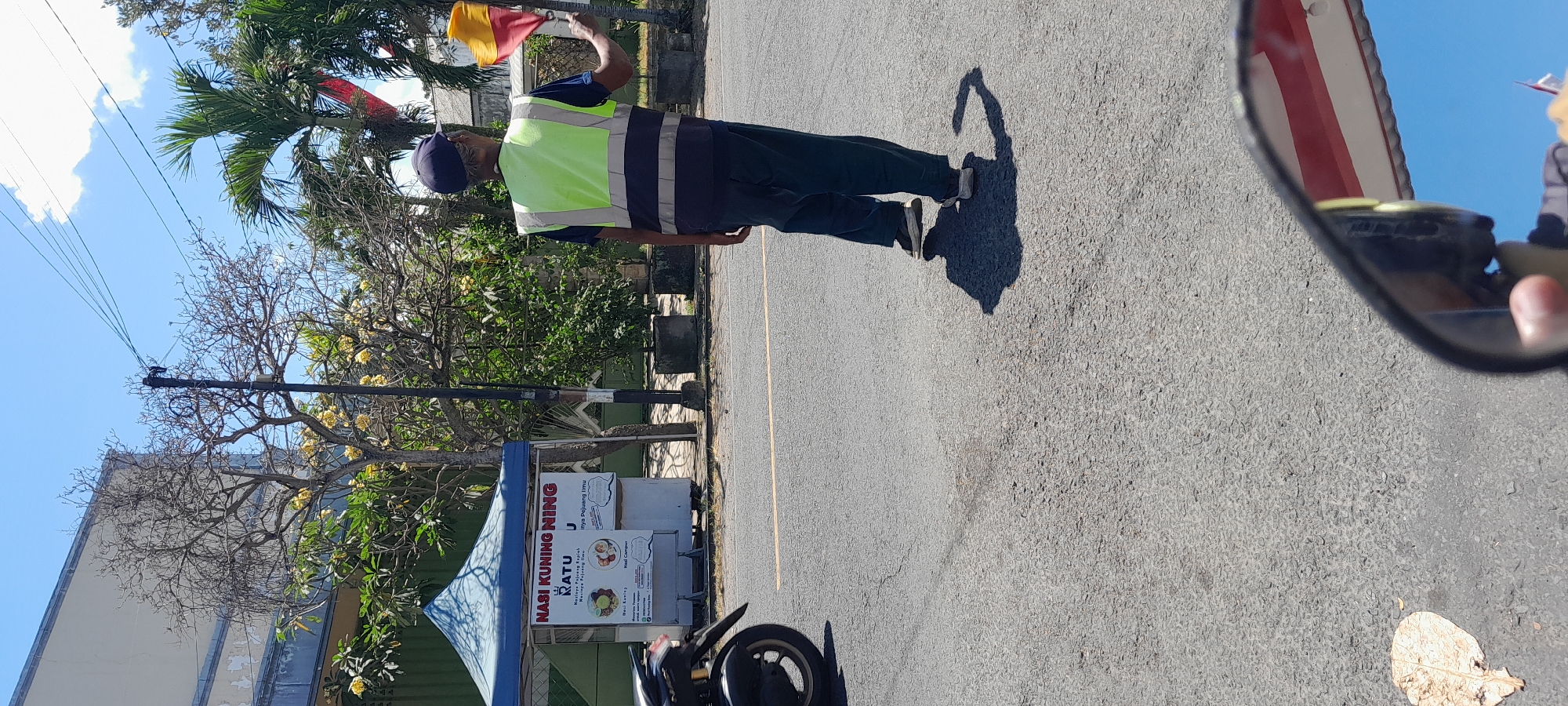 The image depicts a street scene in Rogojampi, Indonesia, under a clear blue sky with scattered clouds. In the foreground, a person wearing a dark blue cap, a neon yellow safety vest with reflective stripes, and dark pants is standing in the middle of the asphalt road, holding two signaling flags, one red and one yellow. The person appears to be directing traffic. To the left, a motorcycle is partially visible, parked on the side of the road. Behind the person, there is a blue tent with signage for "NASI KUNING NING RATU" which appears to be a food stall. Trees with yellow flowers and palm trees are present in the background, along with buildings and power lines. The road has a yellow line painted down the center. A dry leaf is on the asphalt to the lower left, and a side mirror of a vehicle is in the bottom right corner, reflecting the sky and a hint of red and white striped awning.