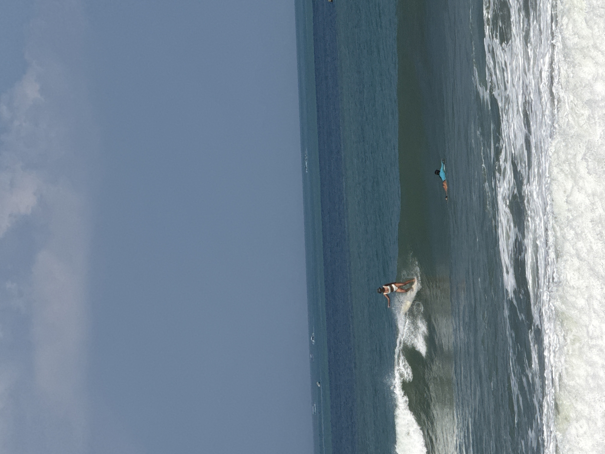 In Kuta, Indonesia, two individuals are engaged in surfing activities in the ocean. In the foreground, a surfer wearing a white bikini and a hat is riding a wave. Further out in the ocean, another surfer is lying on their board, preparing to catch a wave. The scene is set against a backdrop of blue water and a cloudy sky, with several boats visible on the horizon. The image captures a typical day at the beach, showcasing the popular water sport of surfing.